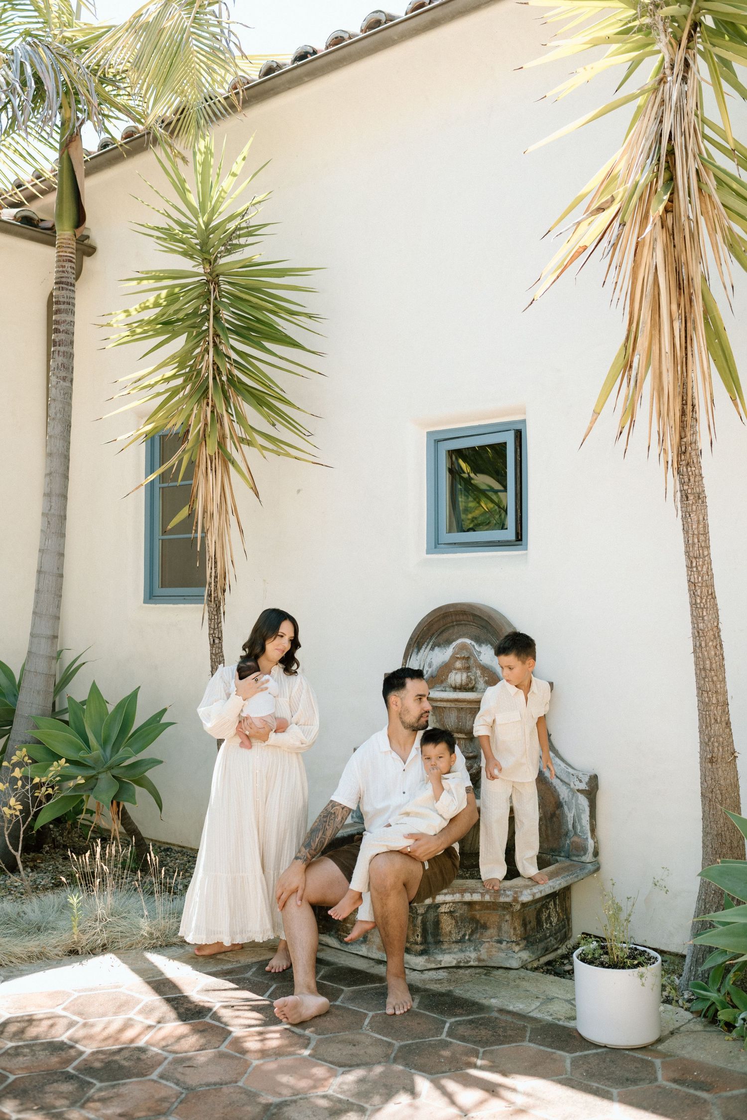 Family poses outside a white stucco home with palm trees, mother holding newborn, father seated with children.