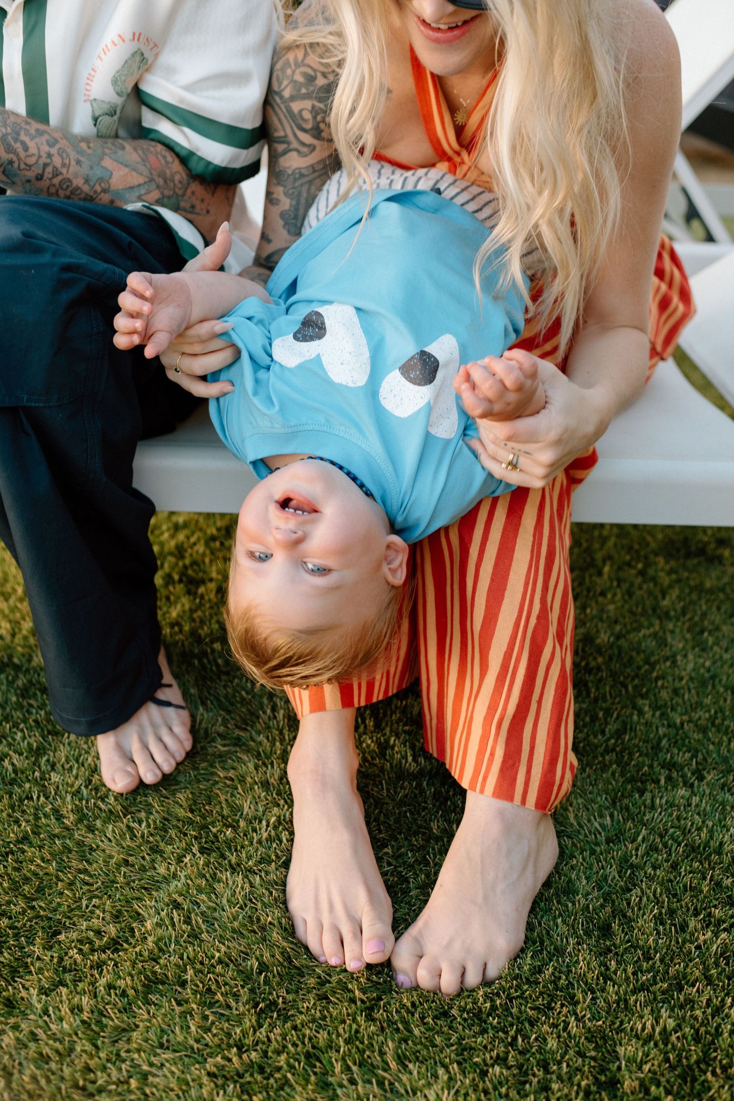 Child wearing a blue monster shirt playfully hangs upside down between two people in a garden setting.