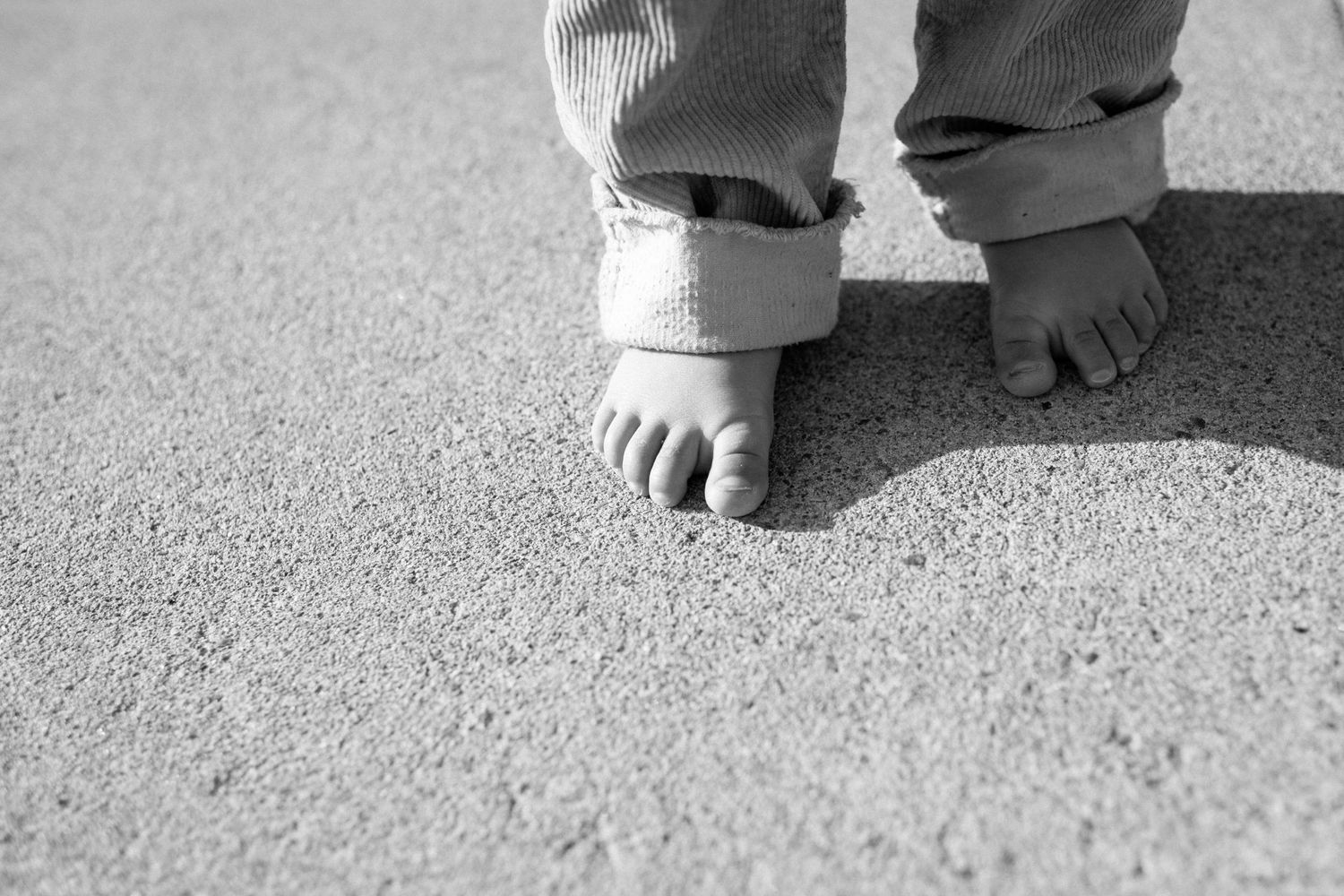 Black and white photo of a toddler's bare feet standing on sandy ground, wearing rolled-up pants.