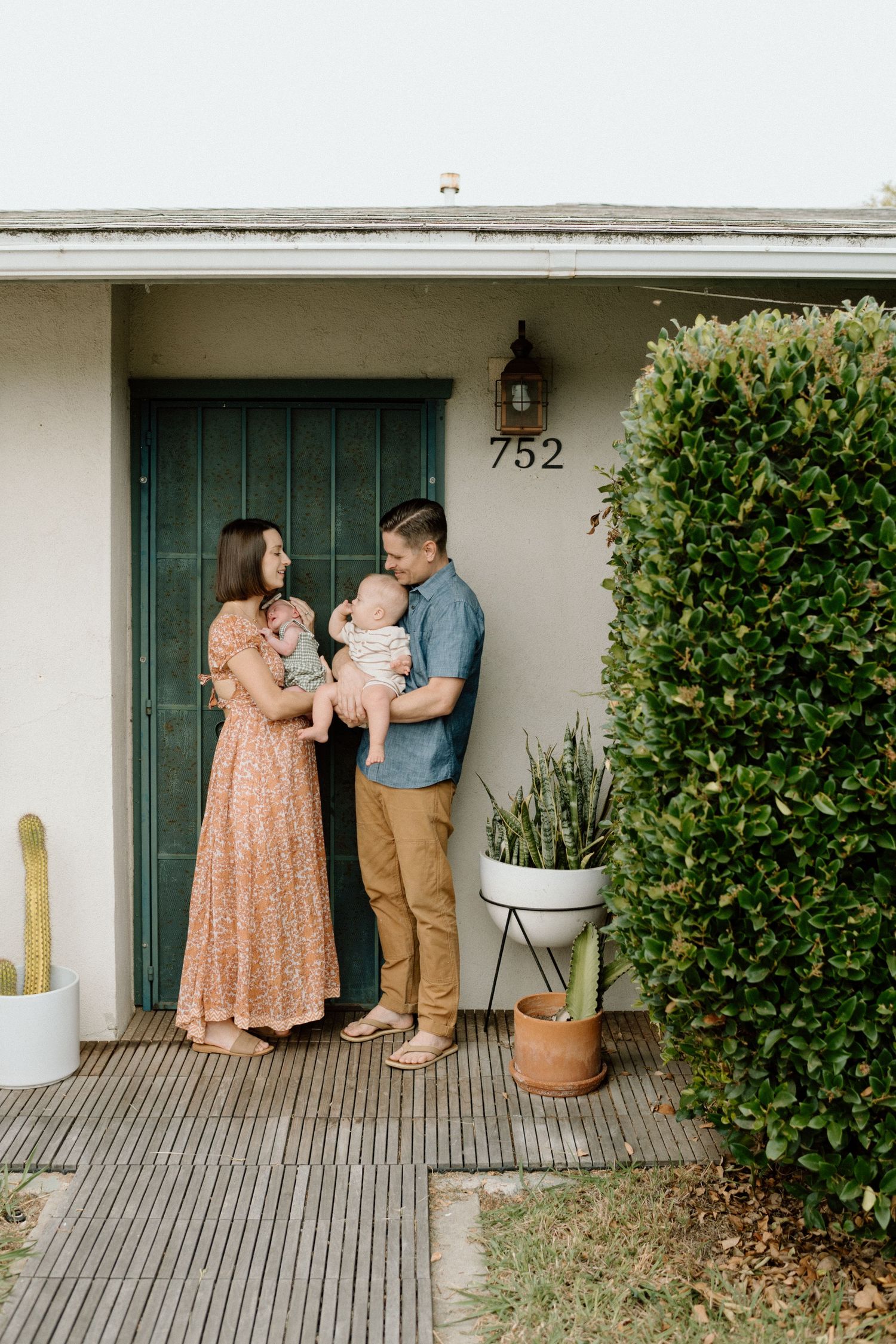 Young family with two babies posing at front door numbered 752, surrounded by green plants and shrubs.