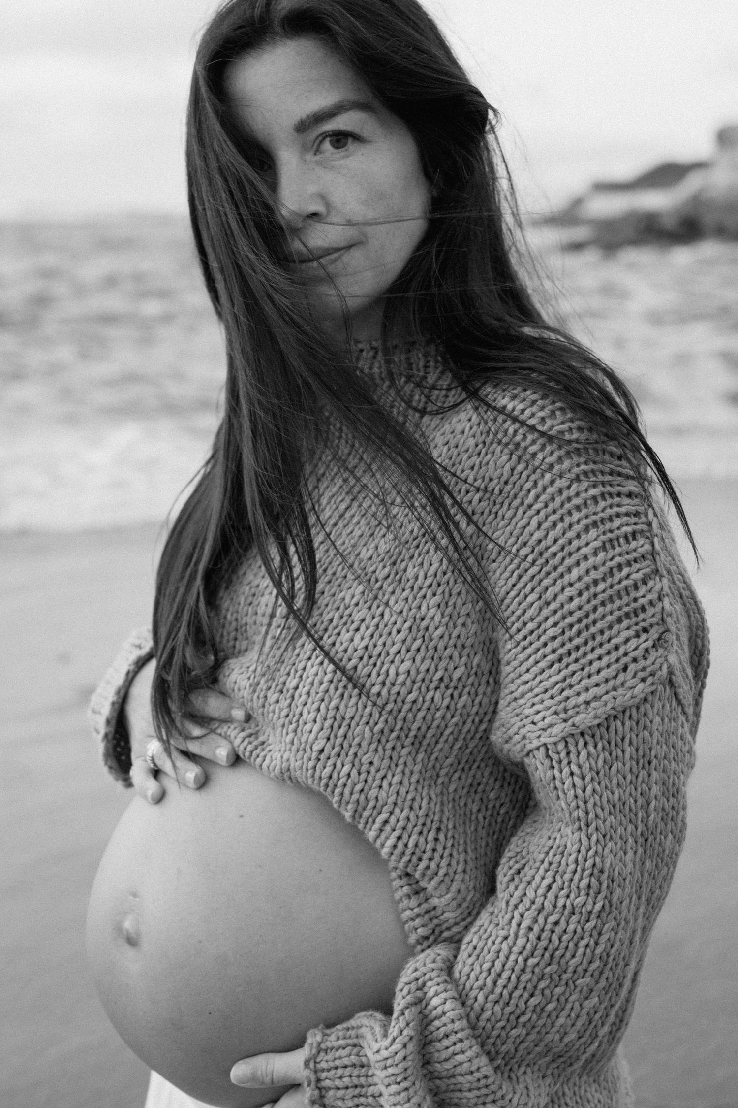 Black and white portrait of pregnant woman in knit sweater at beach.