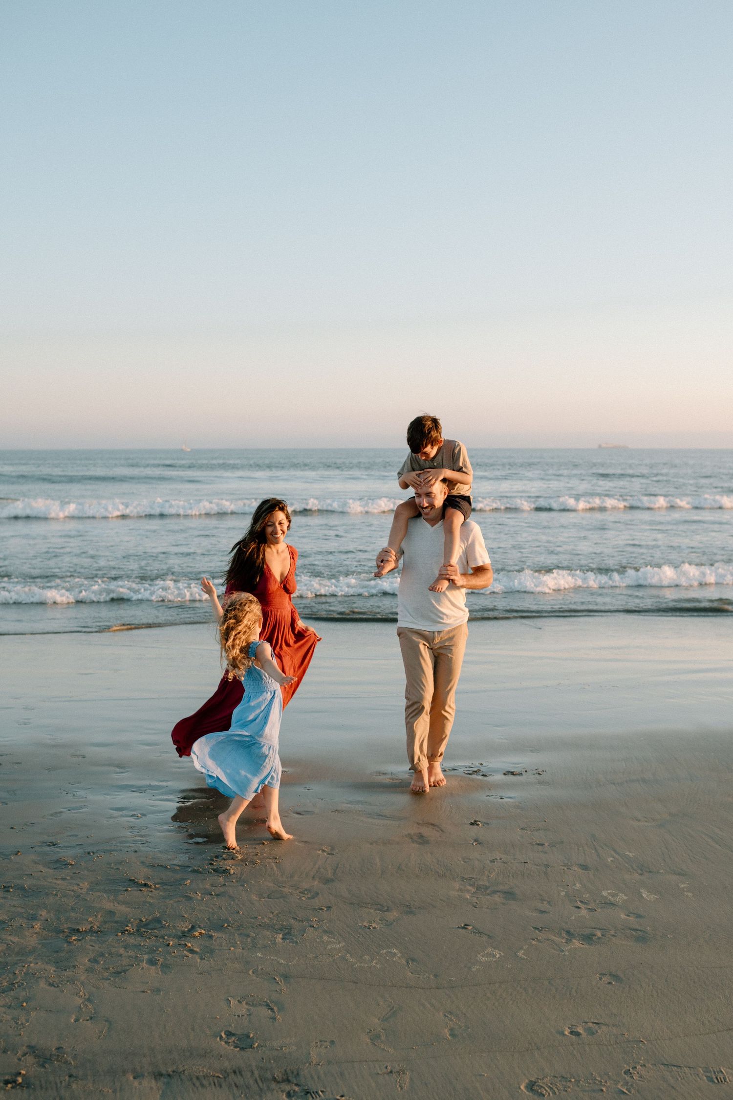 A happy family walks along the ocean shore at sunset, with a child on dad's shoulders.