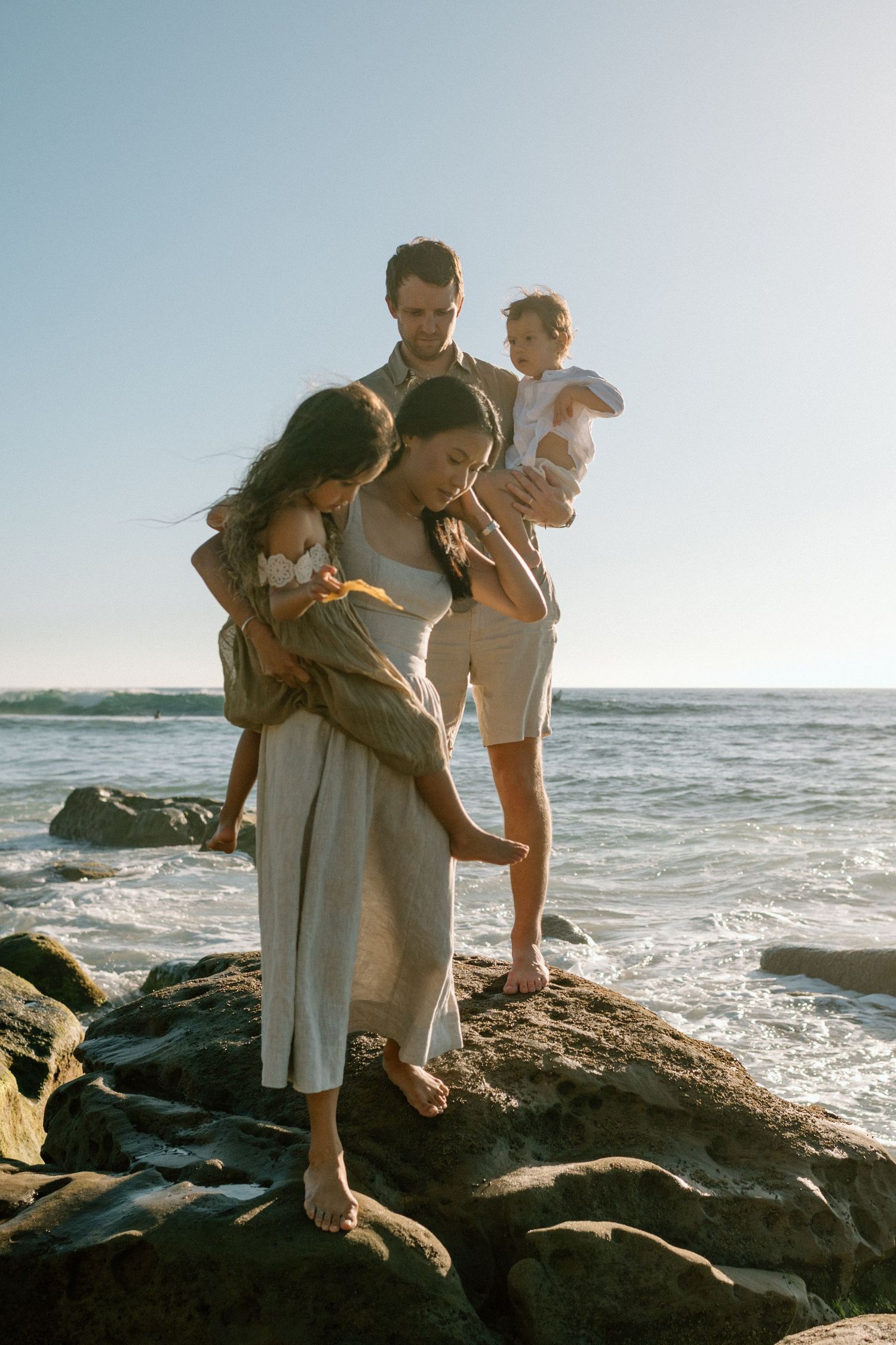 Family of four standing on rocky shore at sunset, parents holding young children with ocean waves in background.