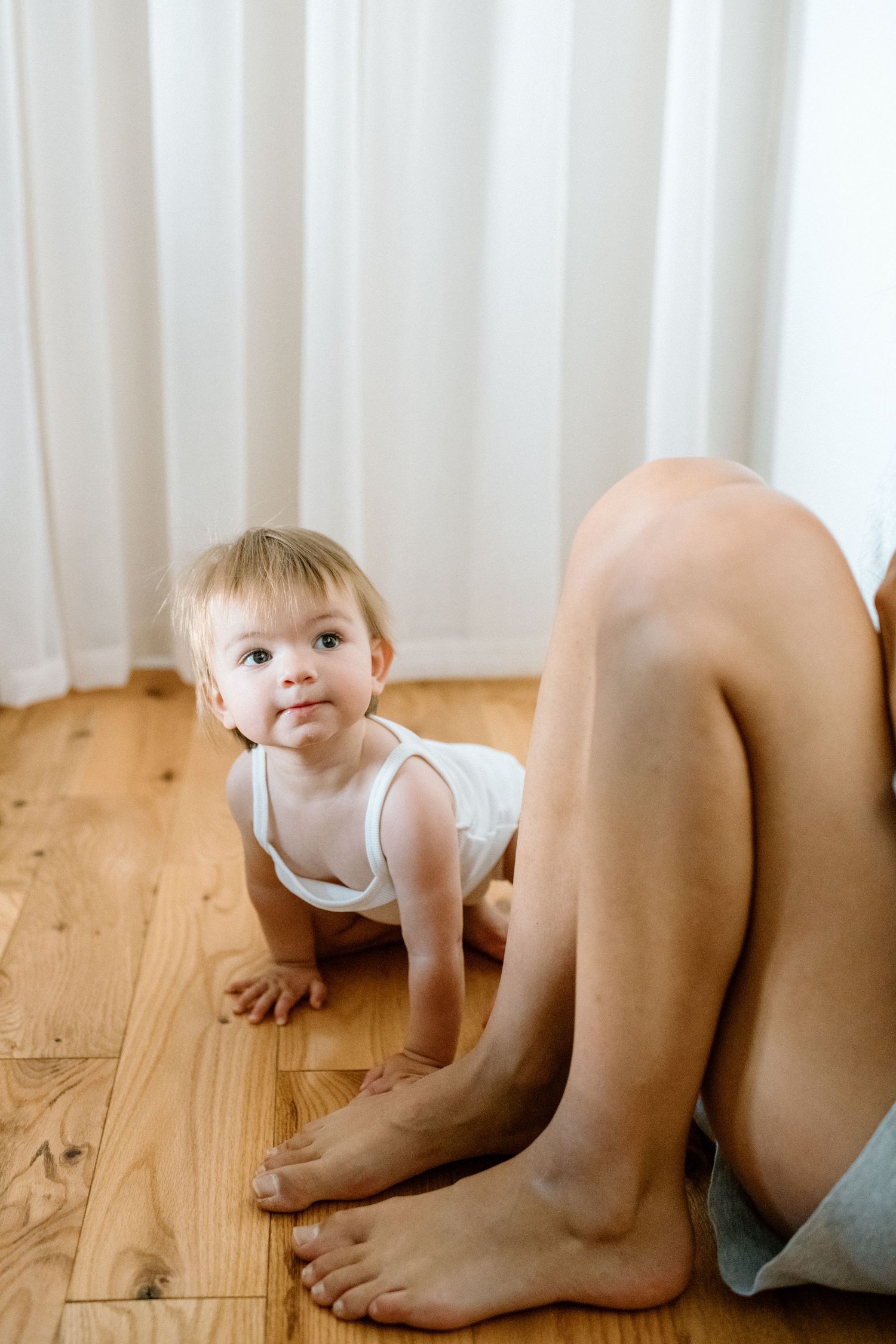 A curious baby in a white onesie crawls on a hardwood floor beside an adult sitting nearby.