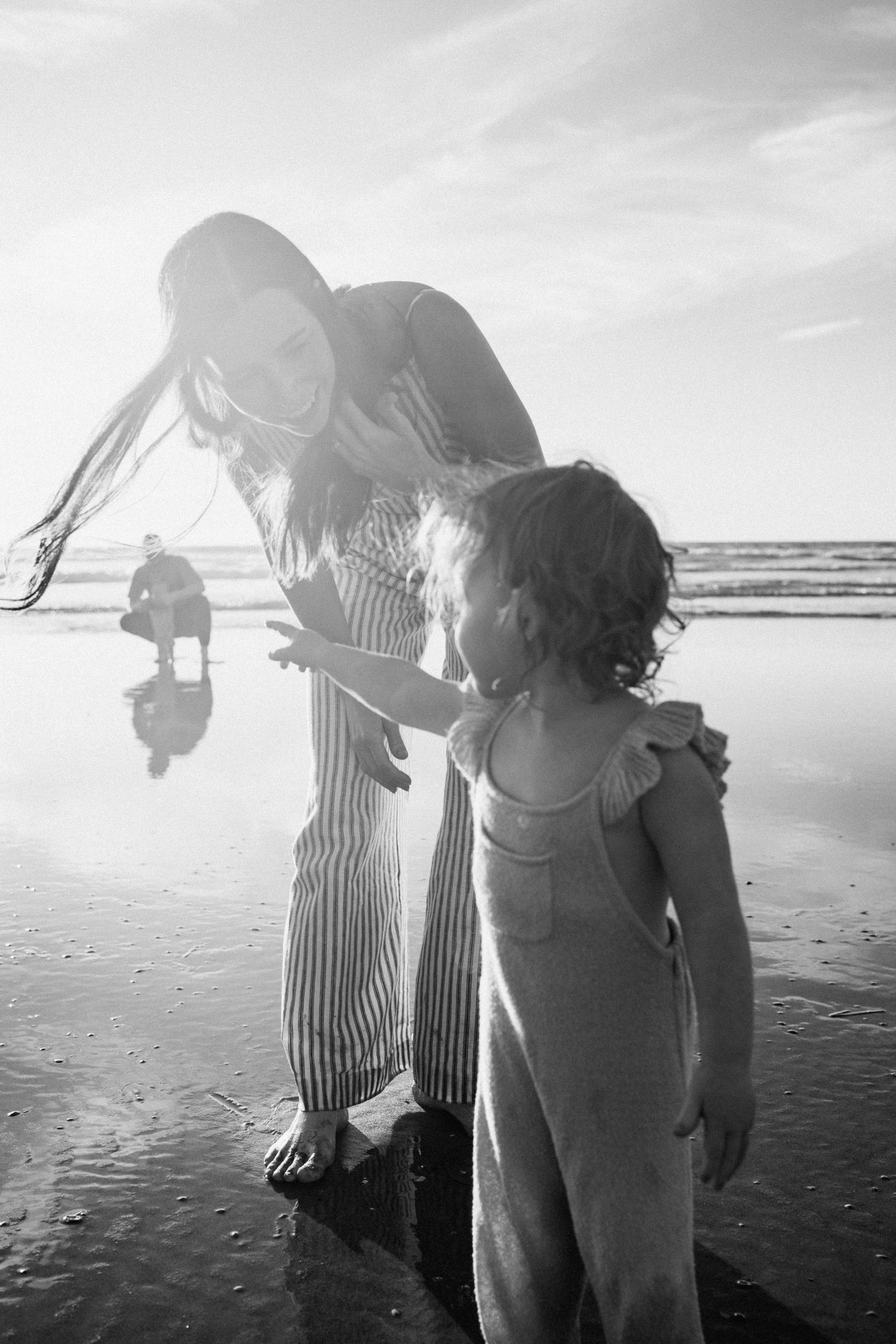 Black and white photo of mother bending down to interact with curly-haired toddler on reflective beach.