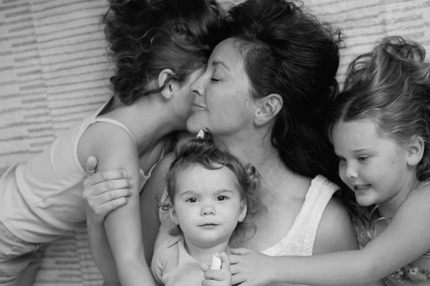 Black and white photo of mother kissing young child while two daughters cuddle close together.