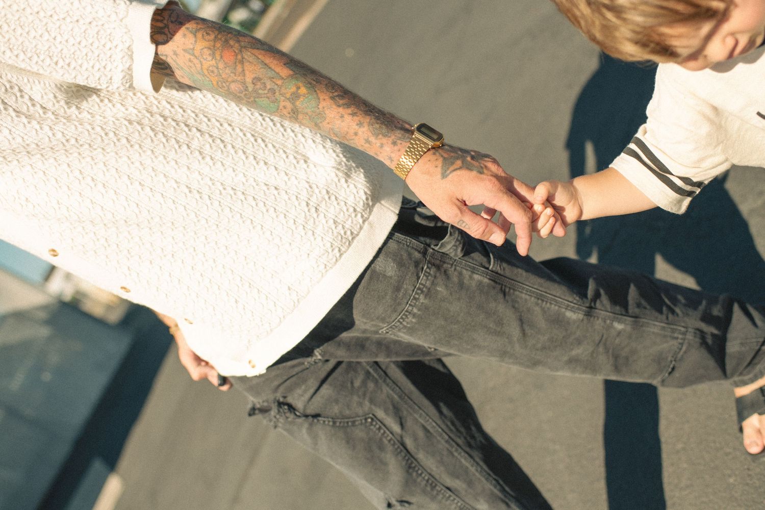 Tattooed adult holding hands with young child while walking outdoors on sunny pavement.
