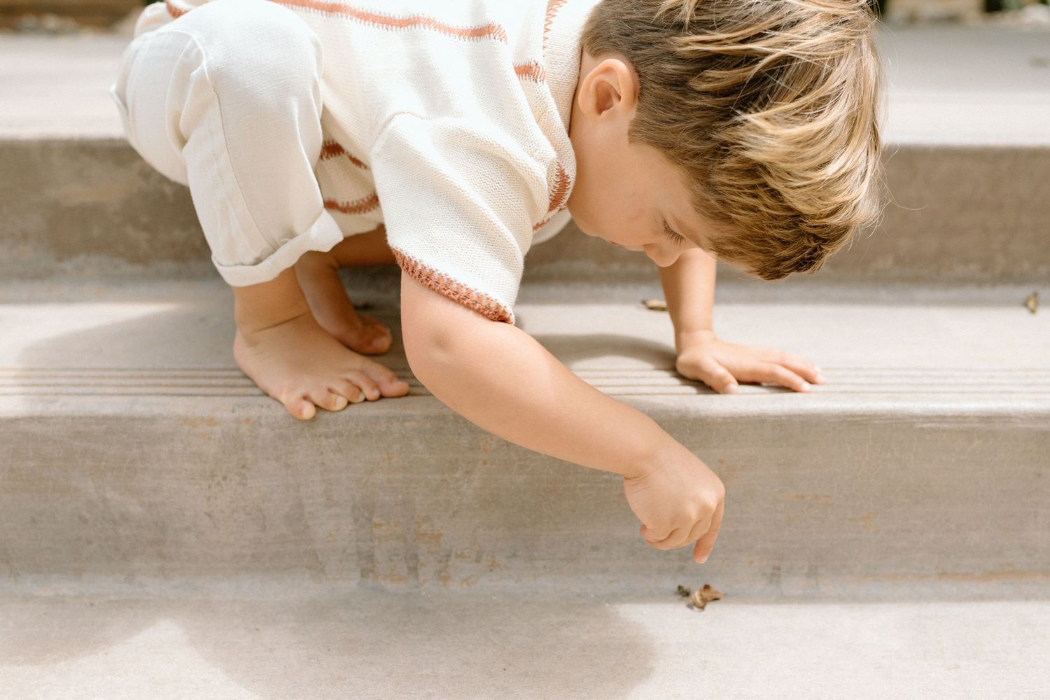 Young barefoot child crouching on concrete steps, reaching down to examine a small object on the ground.