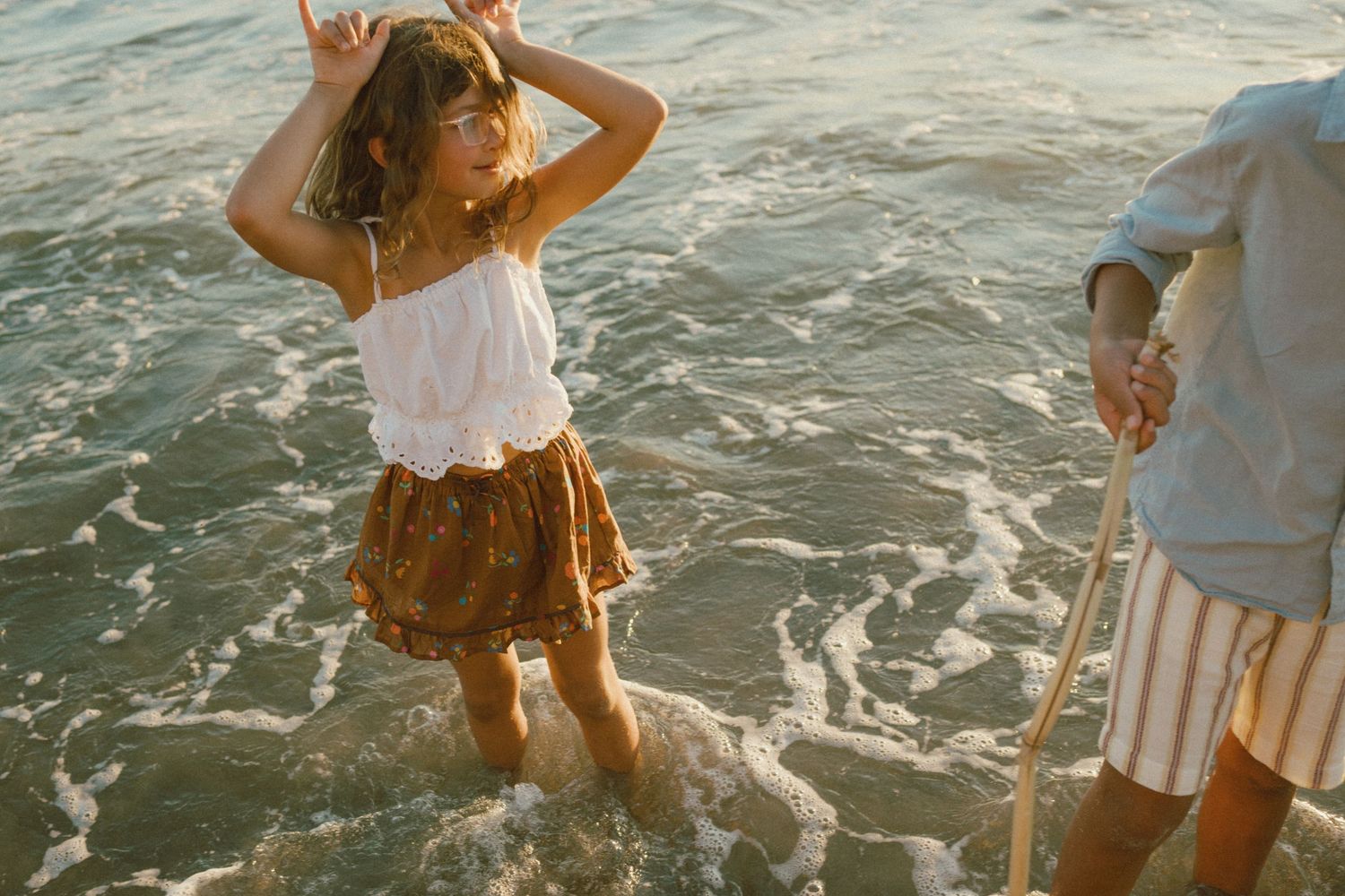 A young girl dances joyfully in ocean waves at sunset, wearing a white top and brown floral skirt on a sandy beach.