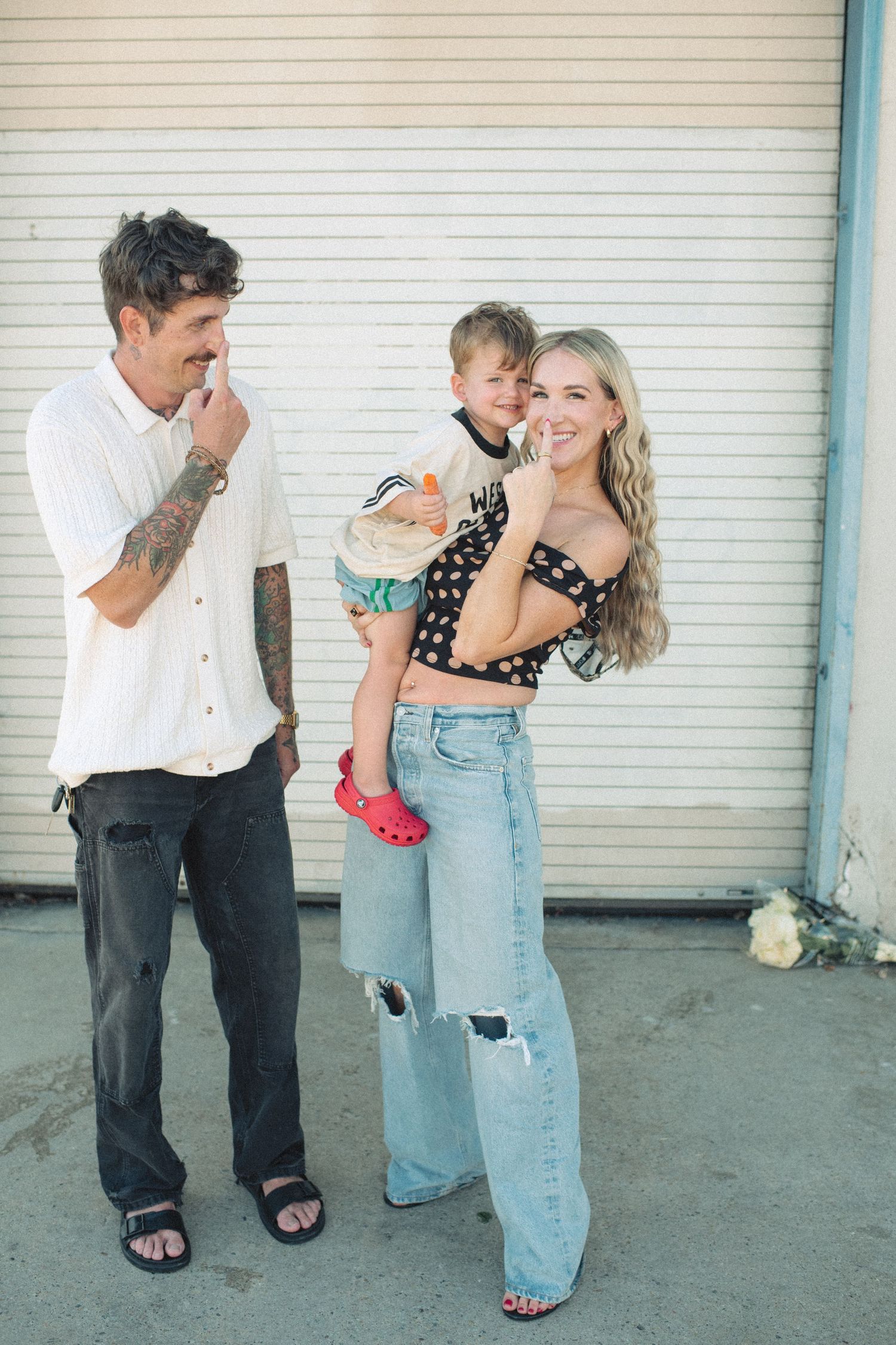 A tattooed man and blonde woman holding a toddler pose together outdoors against a white garage door.