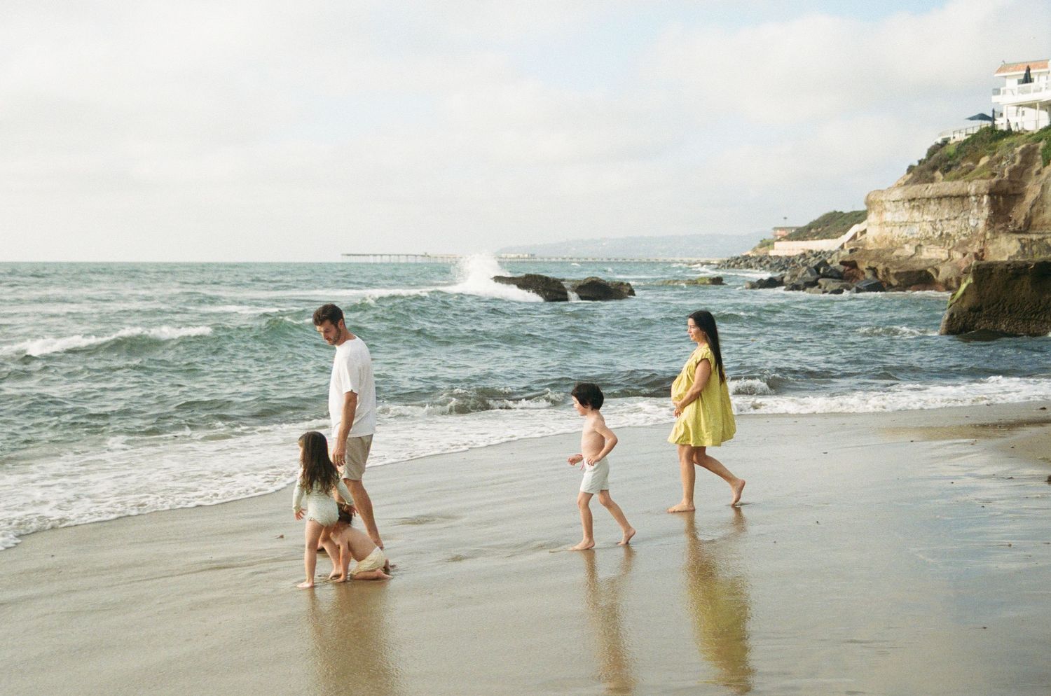 Family of four walking along a sandy beach with ocean waves and rocky cliffs in the background.