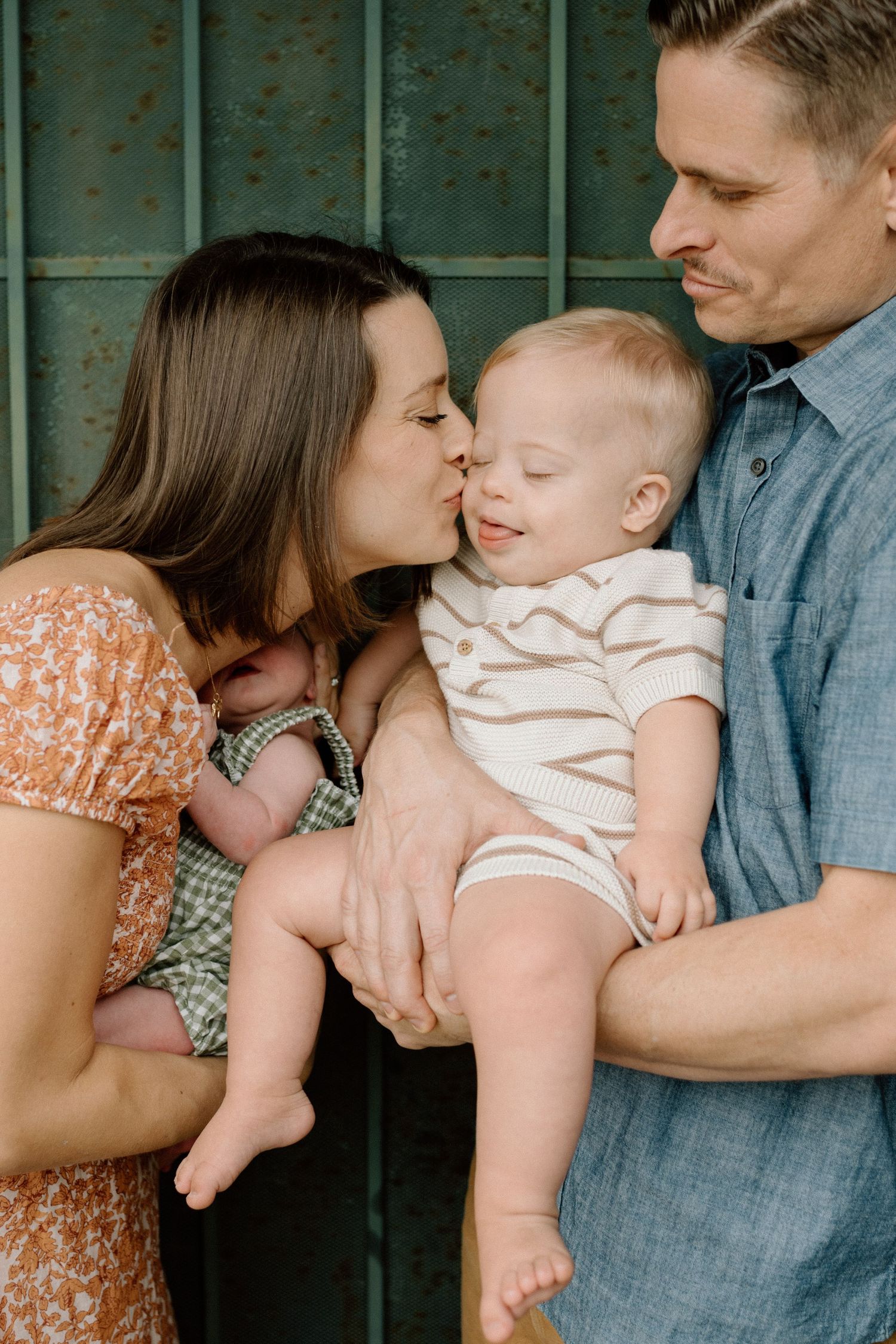 A mother kisses her smiling baby while father looks on, family portrait with two young children.