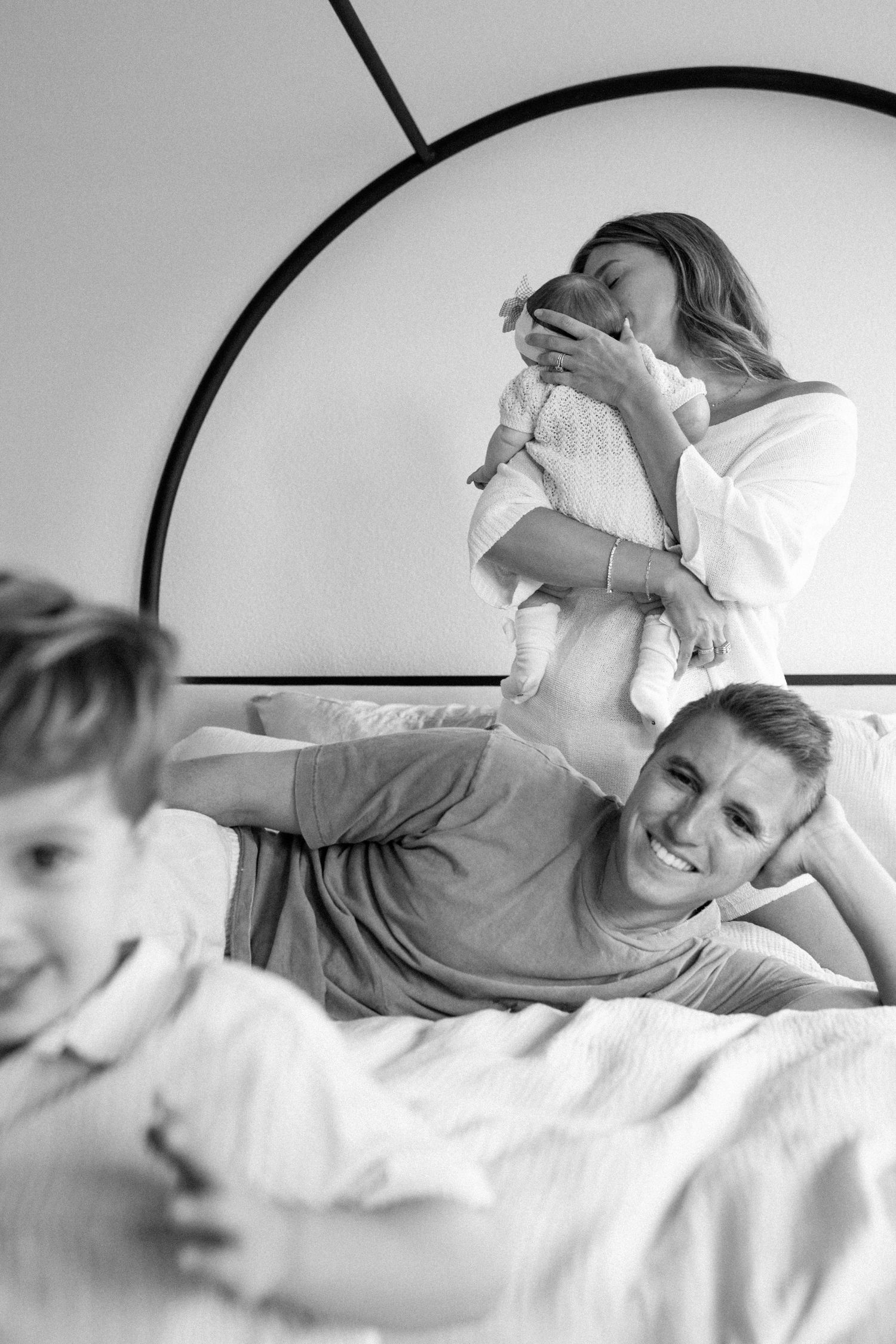 Black and white family photo of mother holding newborn, father smiling on bed, young child playing in foreground.