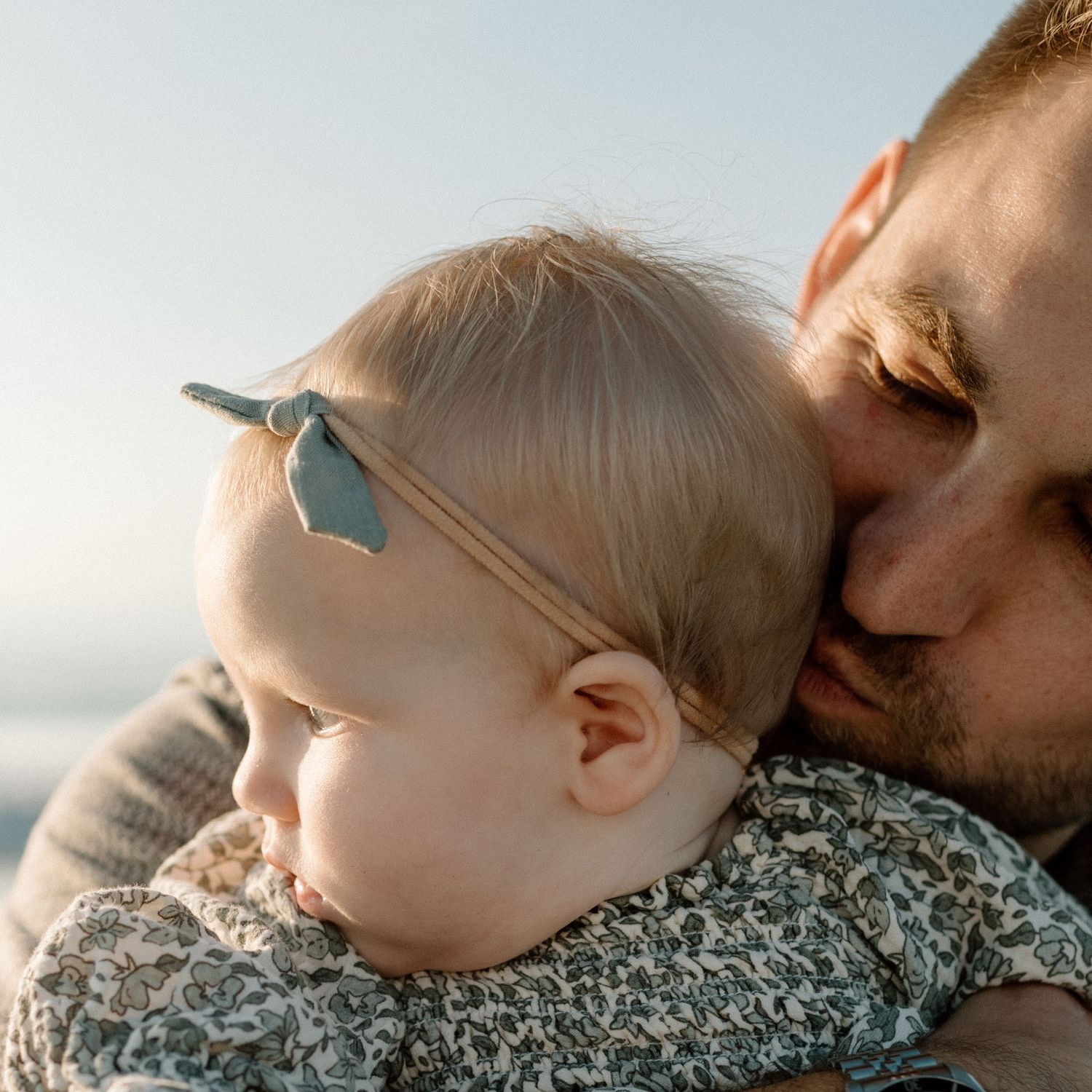 Father tenderly kissing sleeping baby wearing floral outfit outdoors.