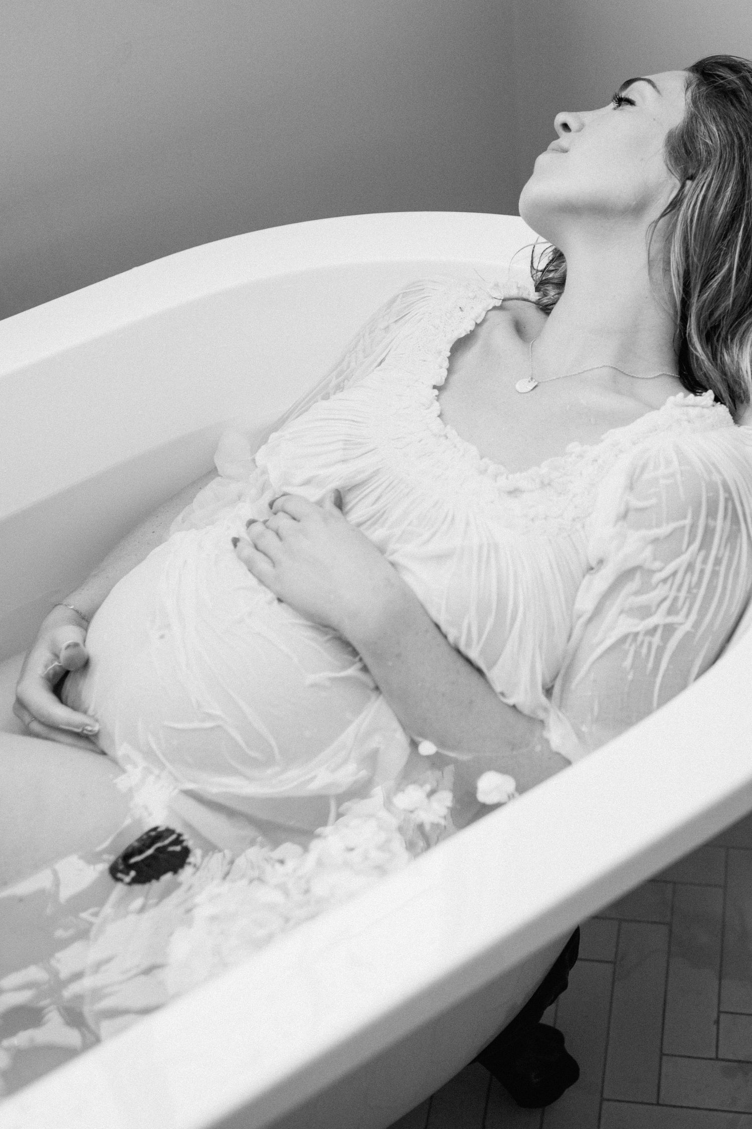 Pregnant woman in white dress reclines in clawfoot bathtub with milk bath and flower petals, black and white photo.
