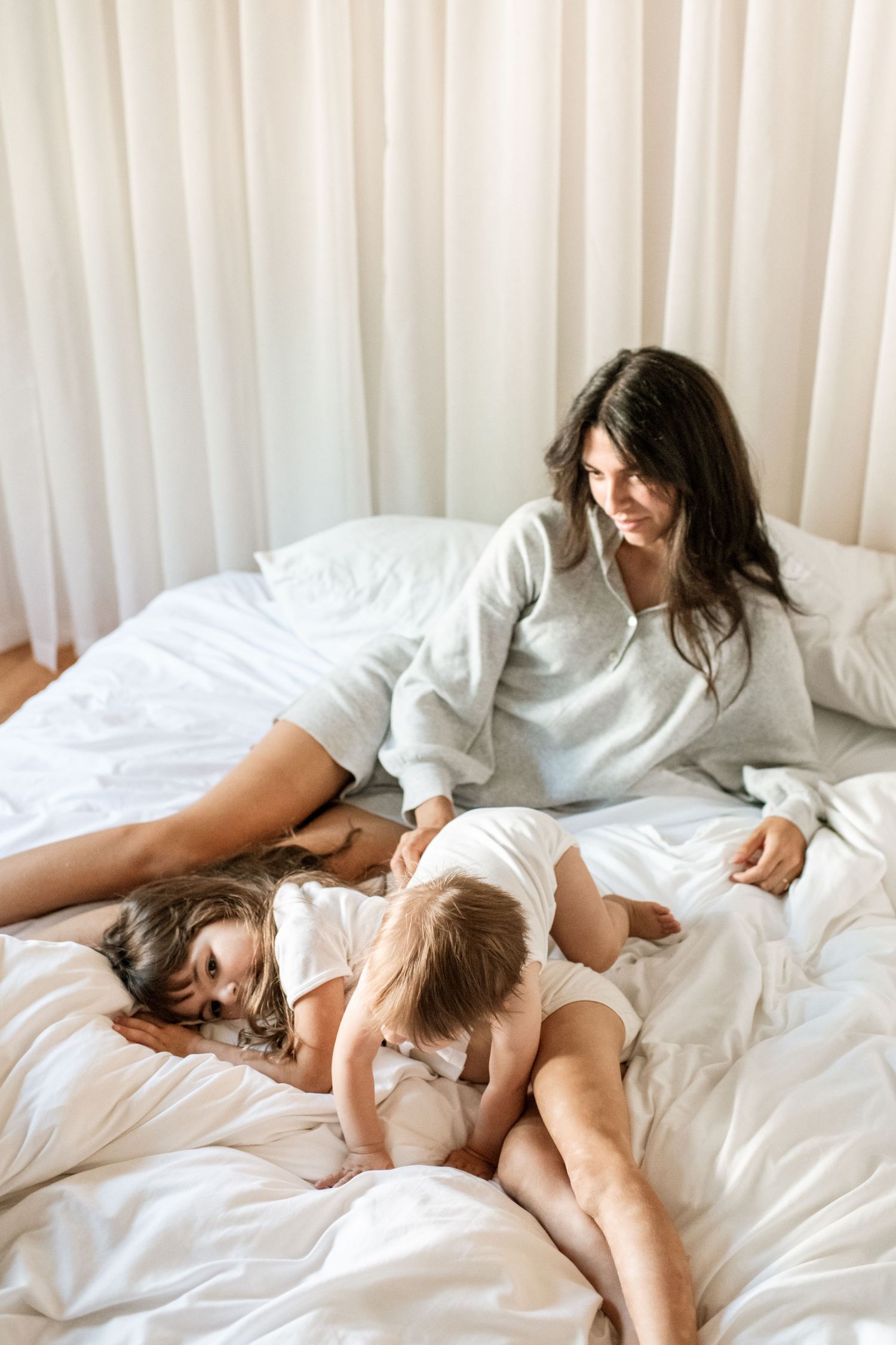 A mother relaxes on a white bed with her two young children in a bright, airy bedroom with sheer curtains.