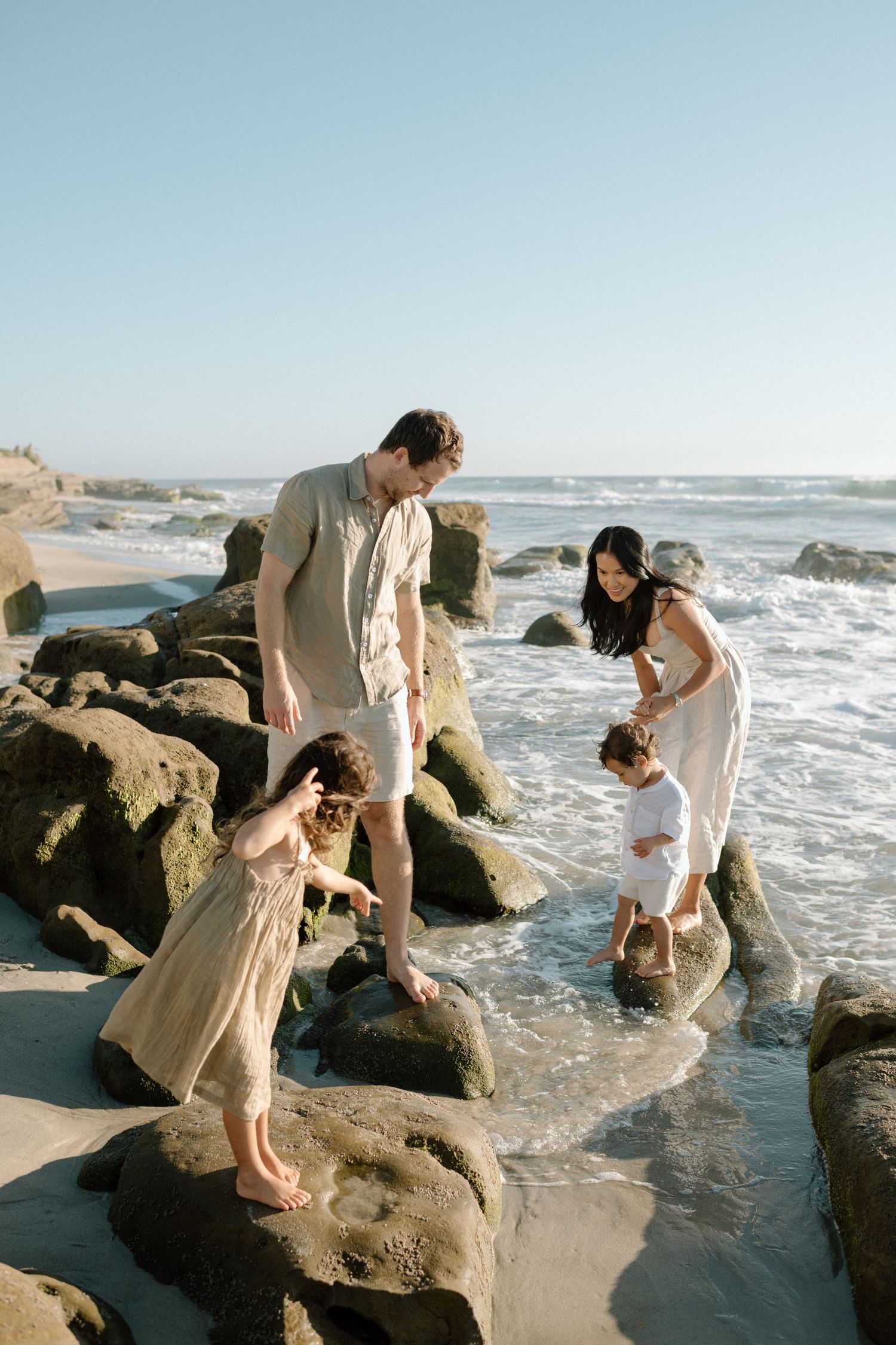 Family of four explores rocky tide pools on a sunny beach with ocean waves in the background.
