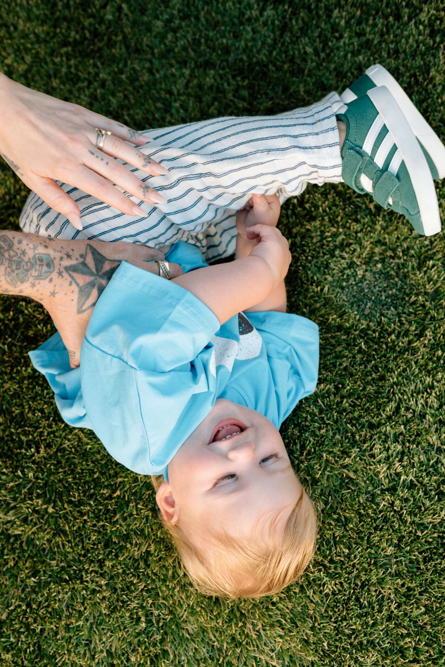 Baby in blue shirt laughs while lying on green grass with a tattooed parent holding their feet.
