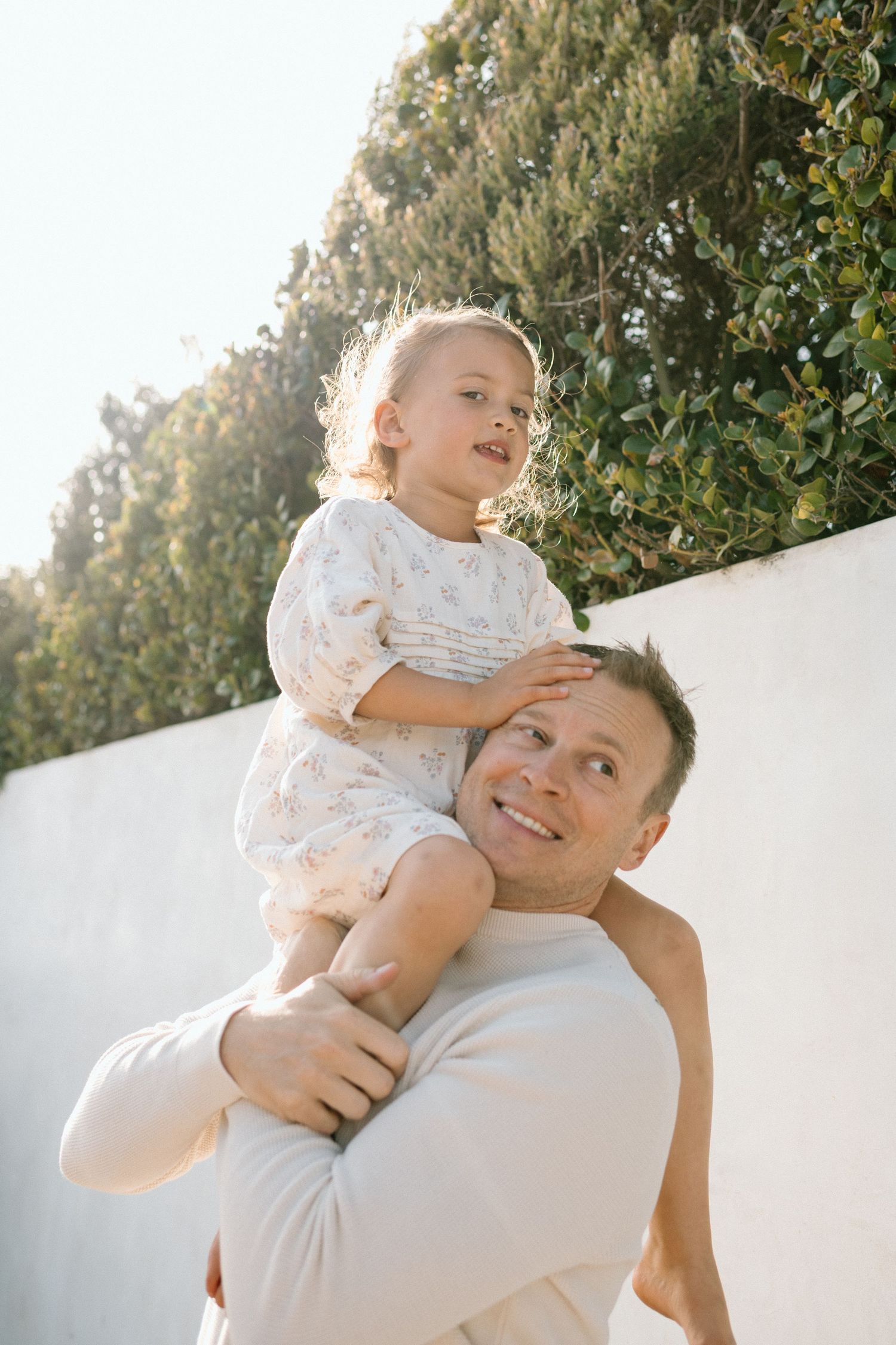 A smiling father carries his young daughter on his shoulders in front of a lush green hedge in warm sunlight.