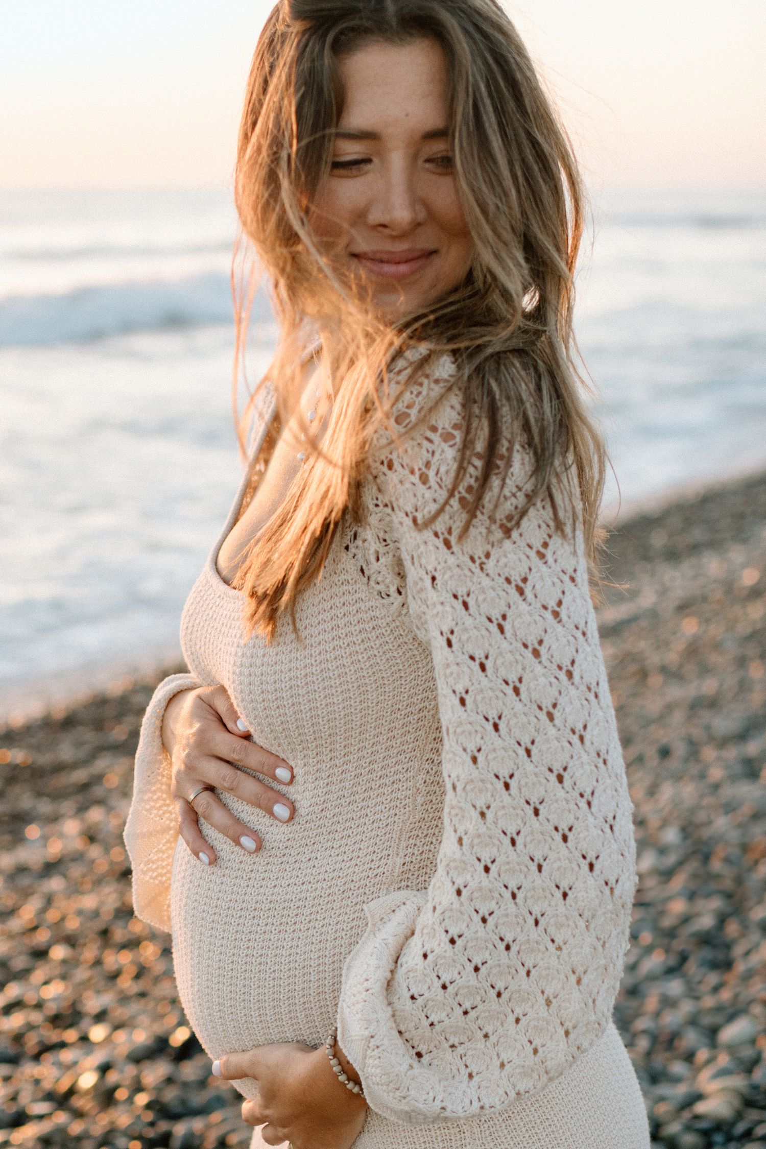 Pregnant woman in cream knit dress cradling her baby bump on a pebble beach at sunset.