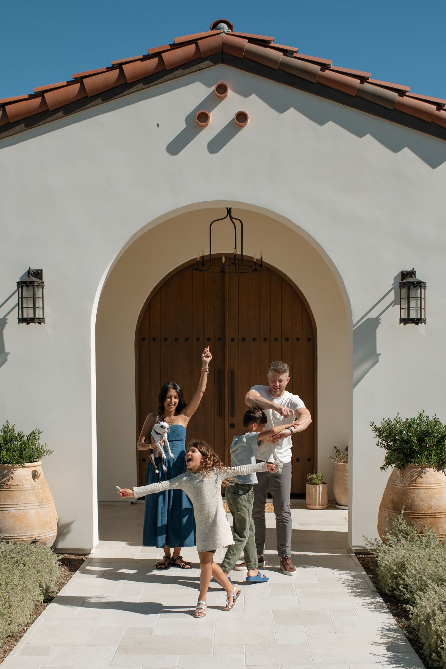 Joyful family of four posing outside elegant white Spanish-style home with arched wooden door.