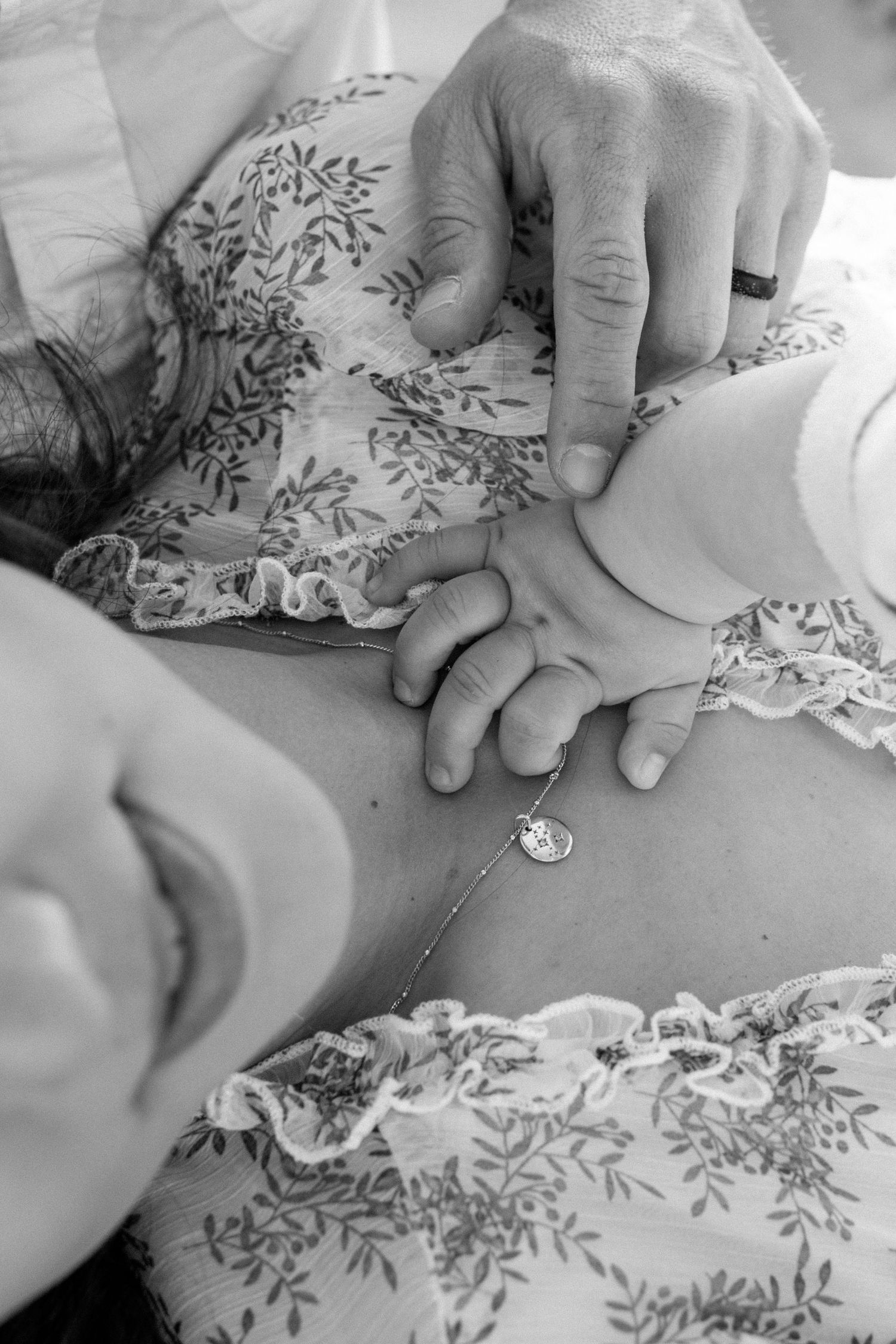 A black and white close-up of a newborn baby holding an adults finger, wearing a floral outfit.