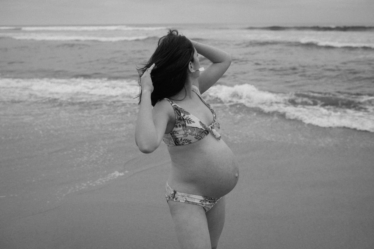 Pregnant woman in patterned bikini standing on beach shore with waves in black and white photo.