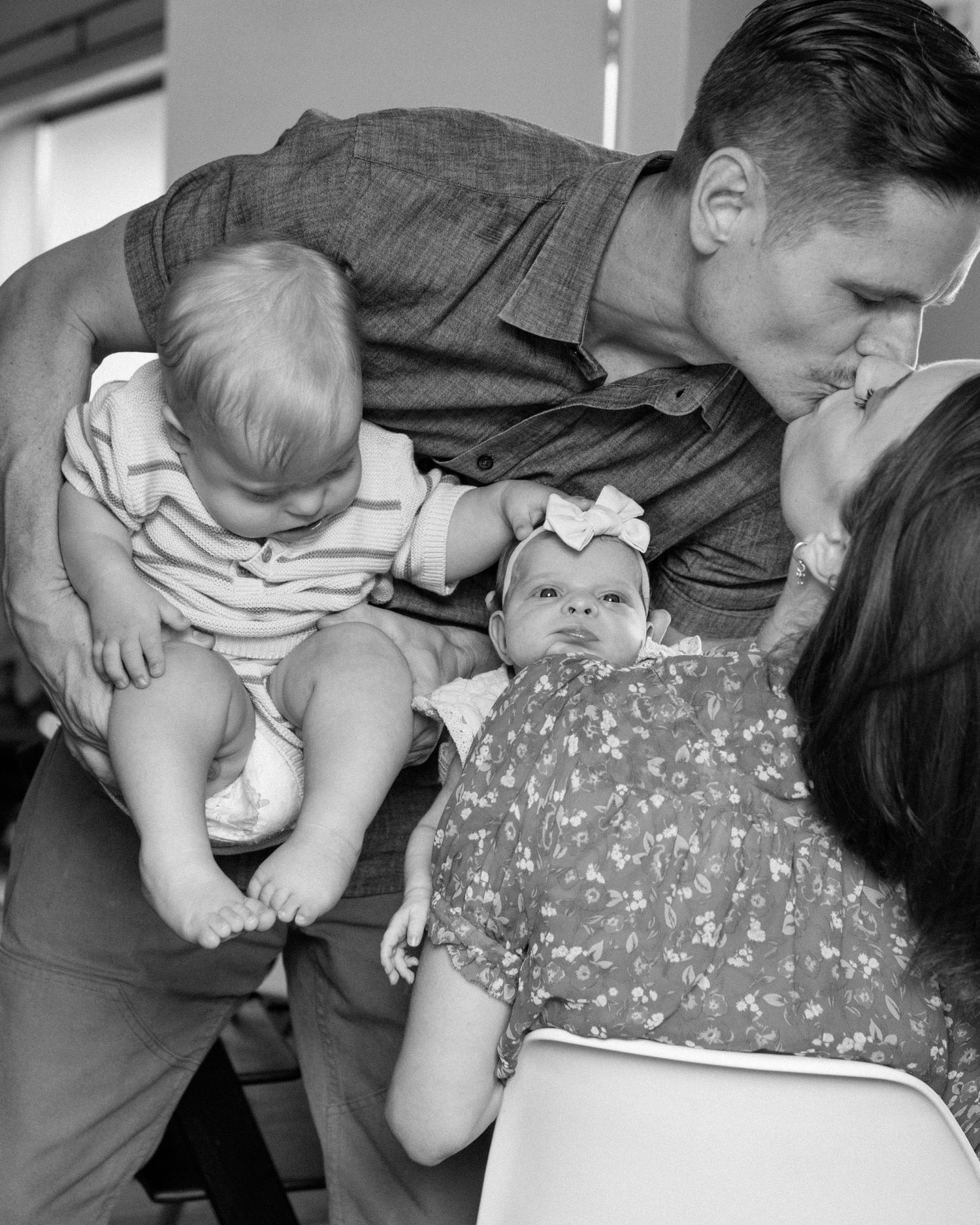Black and white family photo of parents kissing over two babies, one toddler and one newborn girl with a bow.