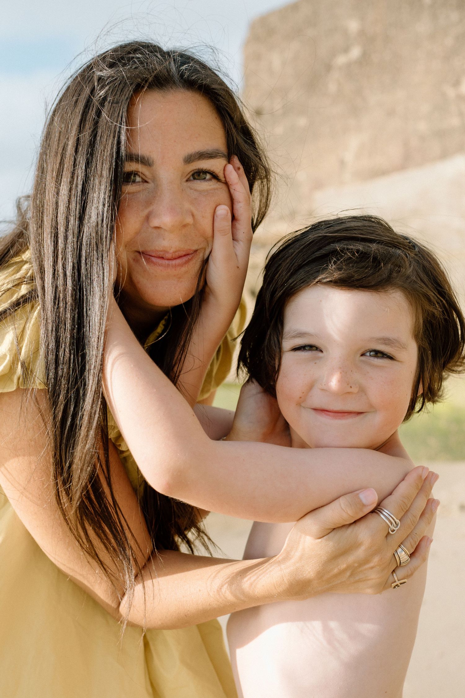 Mother in yellow dress smiling as young child lovingly touches her face outdoors.