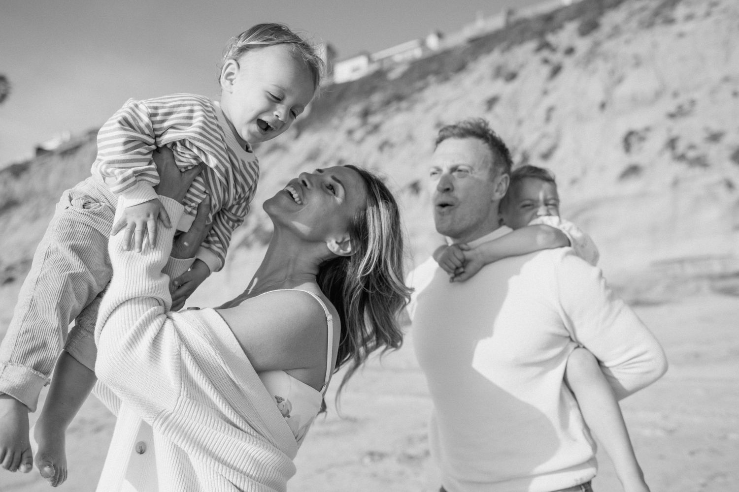 Black and white beach photo of a joyful family with two young children, parents smiling outdoors.