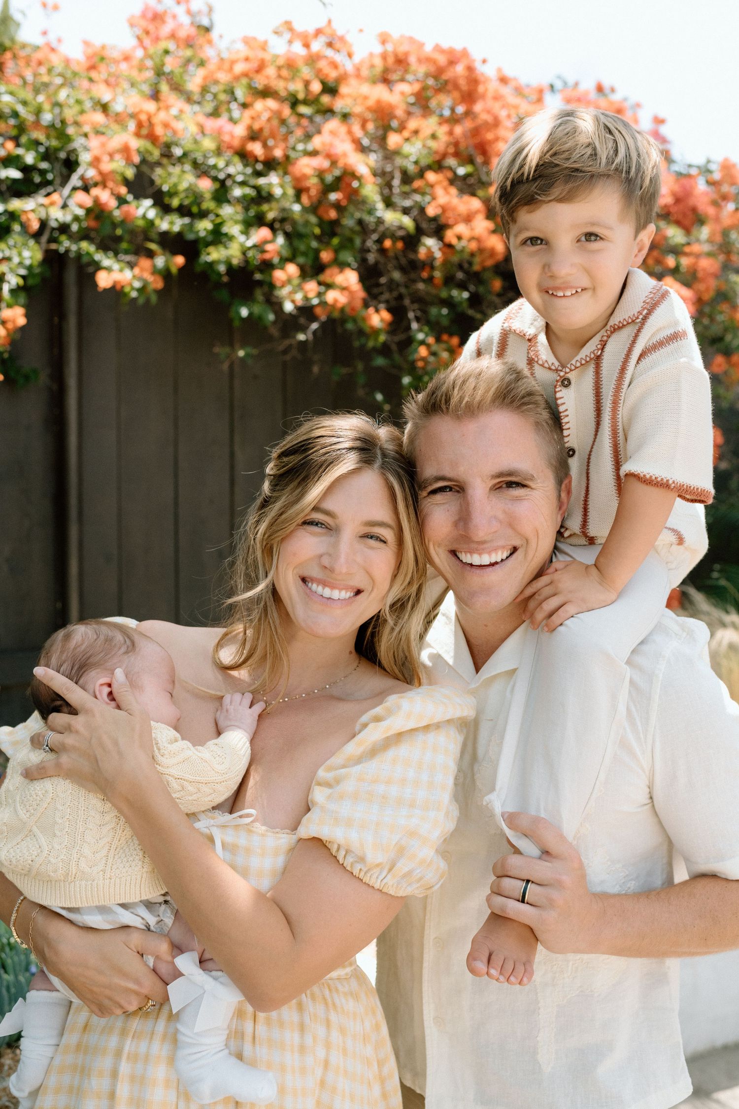 Happy family posing outdoors with newborn baby and toddler son, smiling in front of blooming orange flowers.