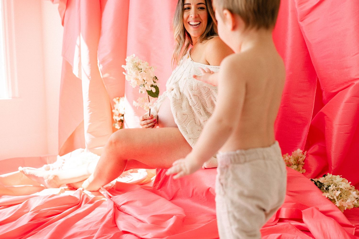 A laughing woman in a white dress sits on pink bedding holding flowers while a toddler in shorts reaches toward her.