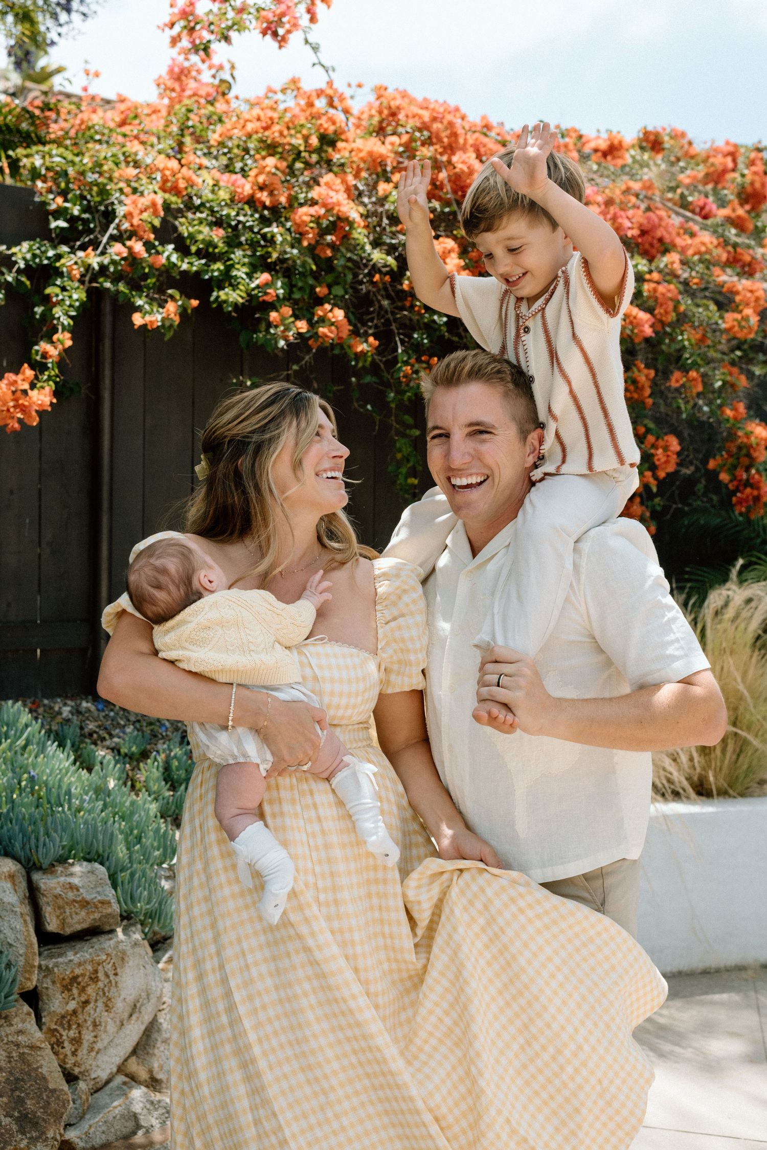 A joyful family laughs together outdoors, with a toddler on dad's shoulders and mom holding a newborn.