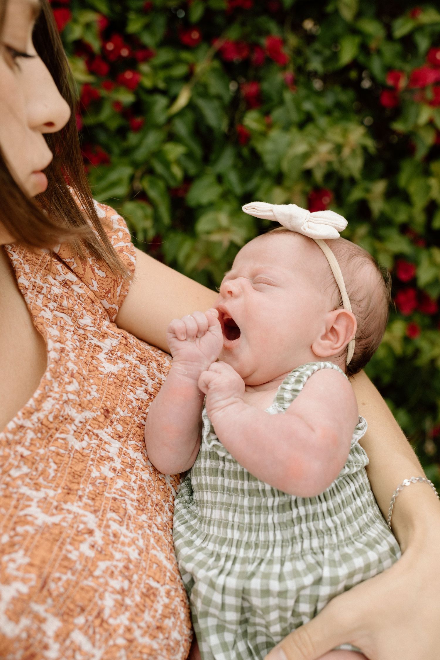 Mother in floral dress holds yawning newborn baby girl in green gingham outfit with white bow headband outdoors.