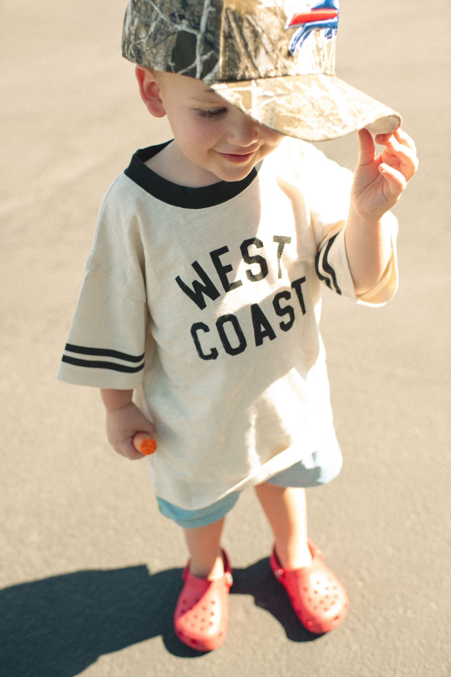 Toddler boy wearing a West Coast tee, camo cap, and red Crocs, standing outdoors in sunlight.