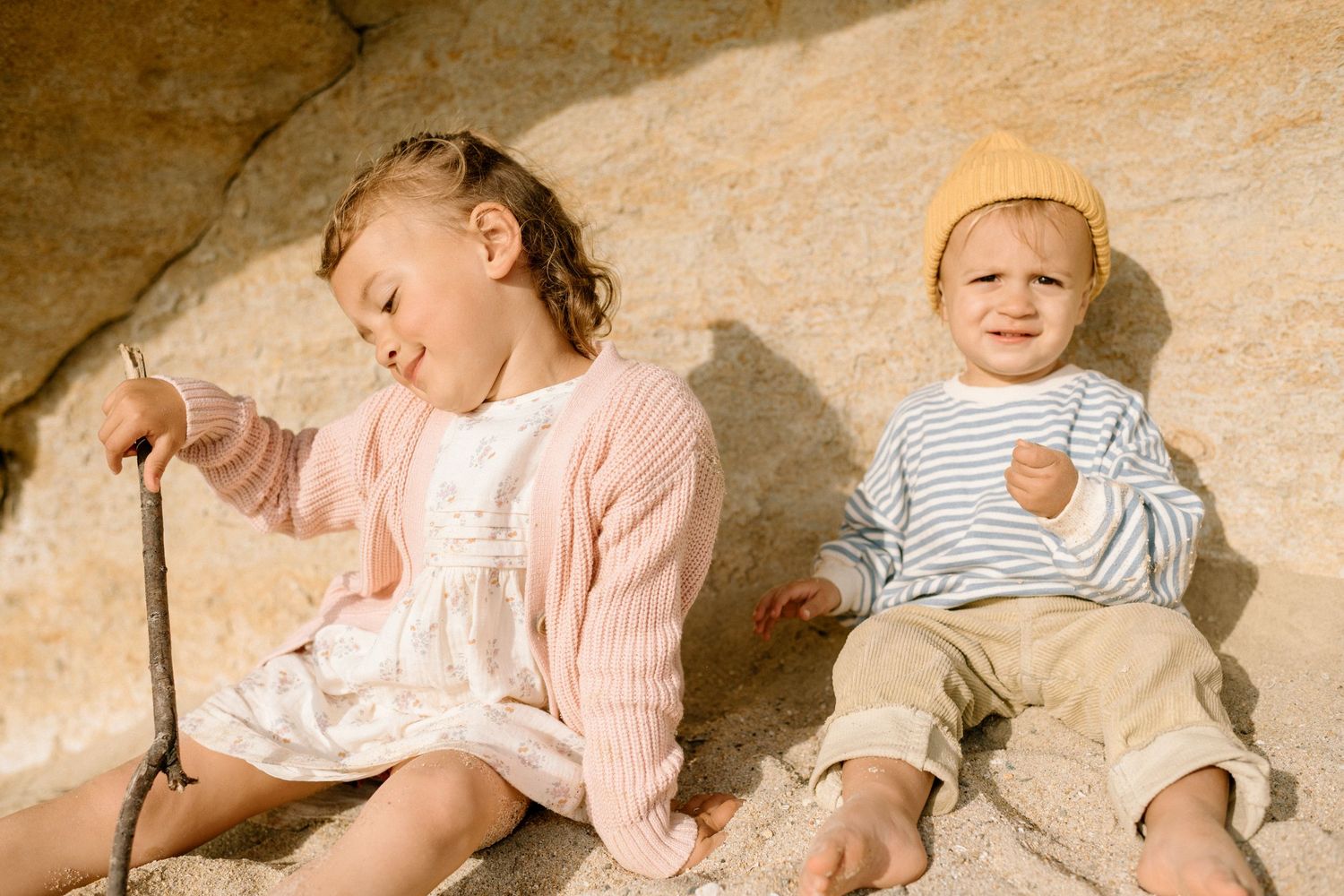 Two young children sitting on sandy rocks, girl in pink cardigan and boy in striped sweater with yellow hat.