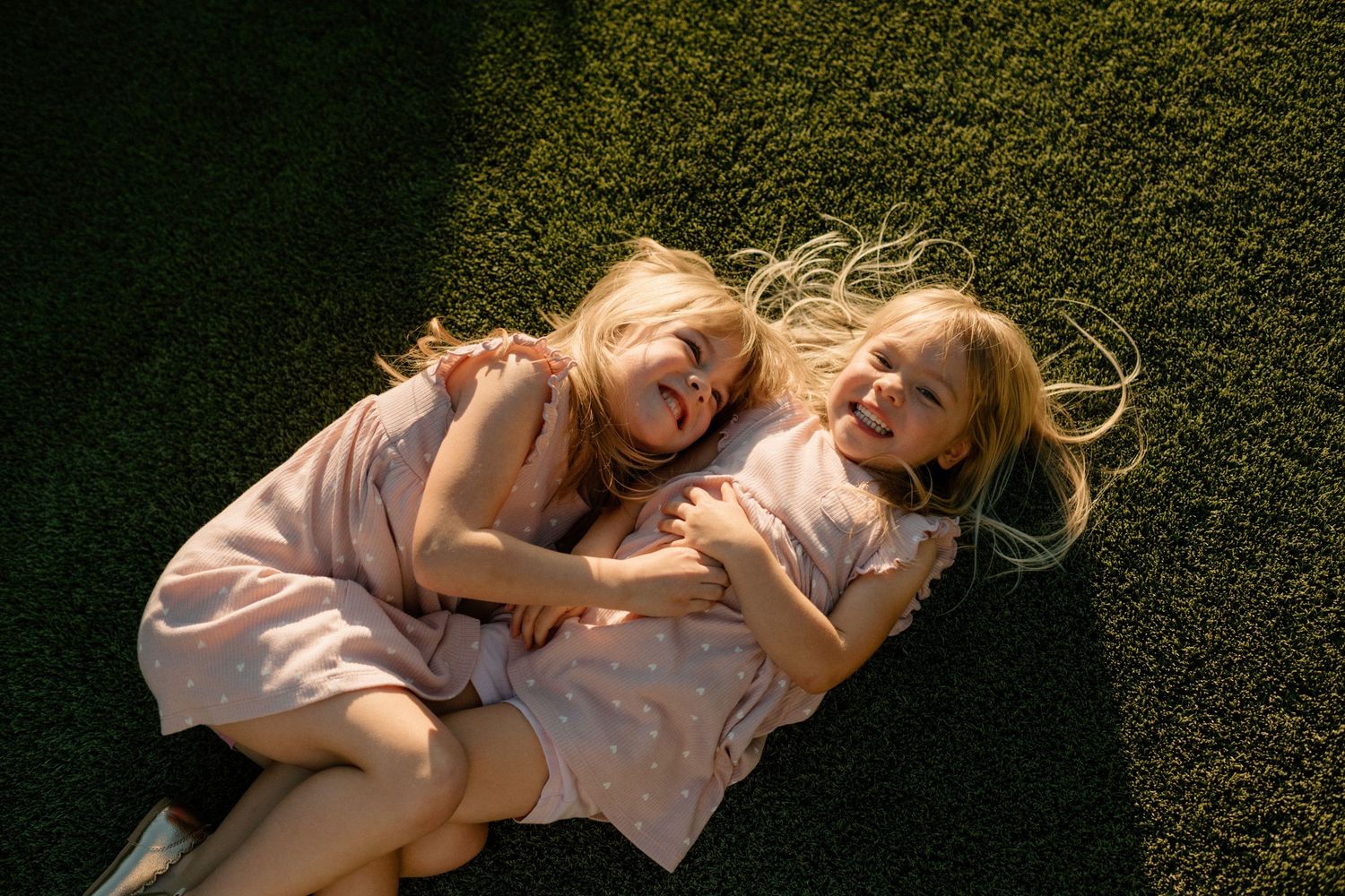 Two blonde girls in matching pink dresses lying on green grass laughing and hugging outdoors.