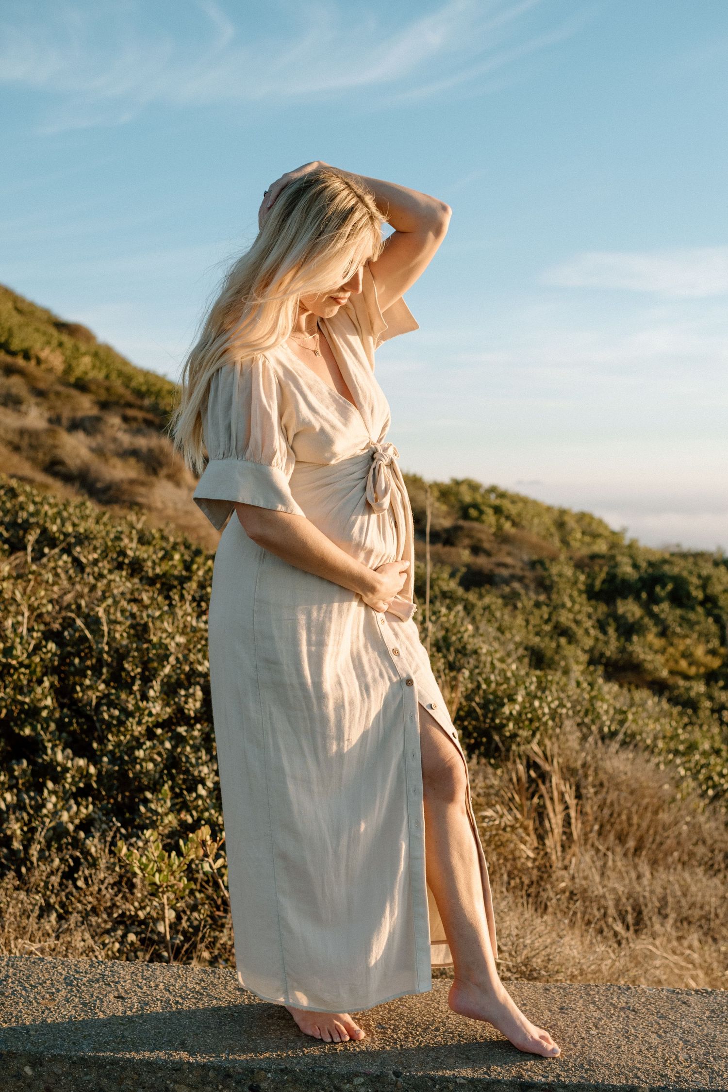 Pregnant blonde woman in white maxi dress cradles bump barefoot on path at golden hour outdoors.