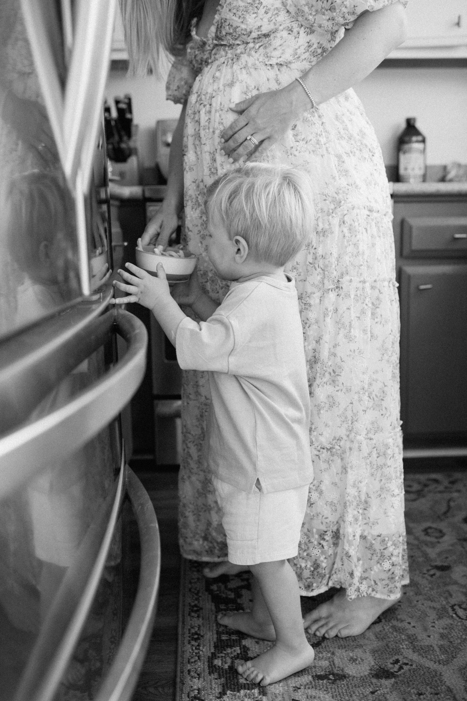 Black and white photo of toddler boy standing beside pregnant mother in floral dress in kitchen.