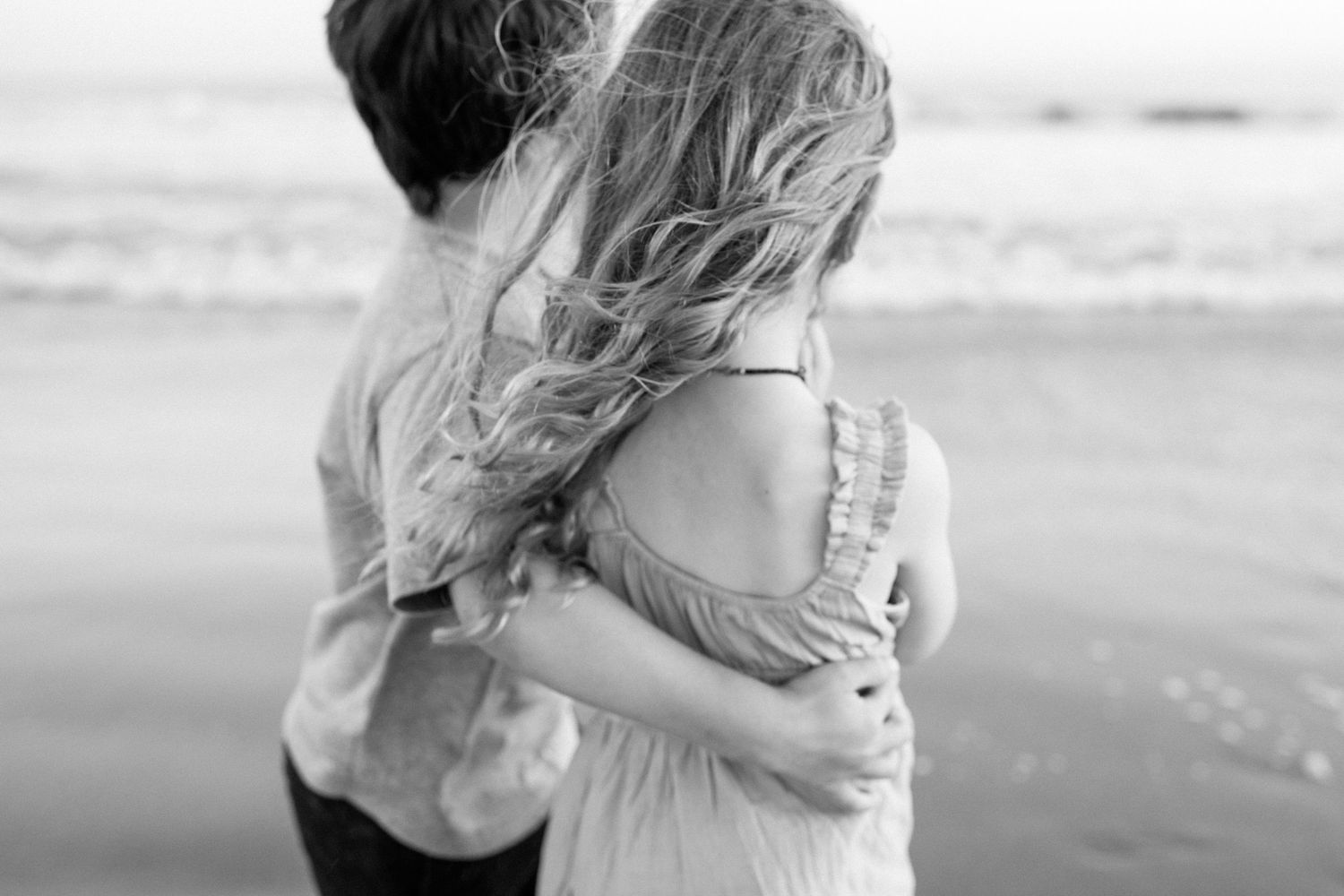 Black and white photo of two children sharing a tender embrace on a breezy beach.