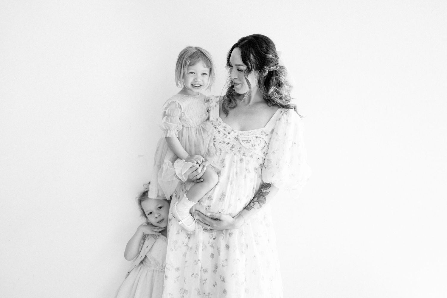 Black and white photo of a pregnant mother in a lace dress holding her bump with two young daughters.
