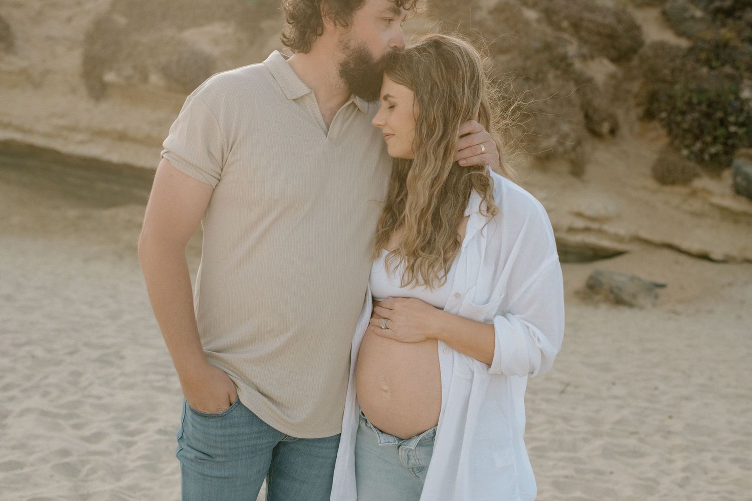 Couple sharing tender moment on beach, pregnant woman cradling baby bump while partner embraces her.
