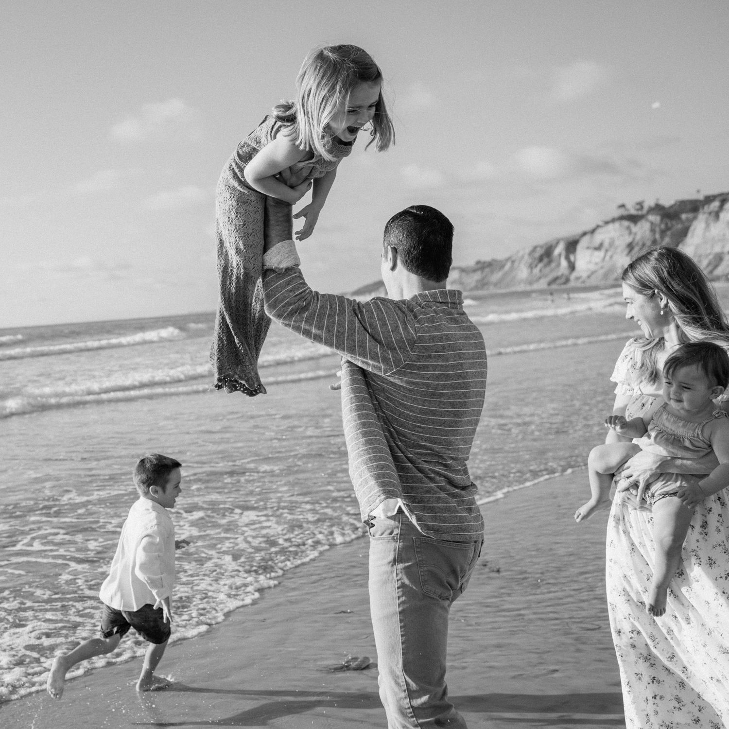 Black and white photo of father lifting daughter on beach while family plays by ocean.