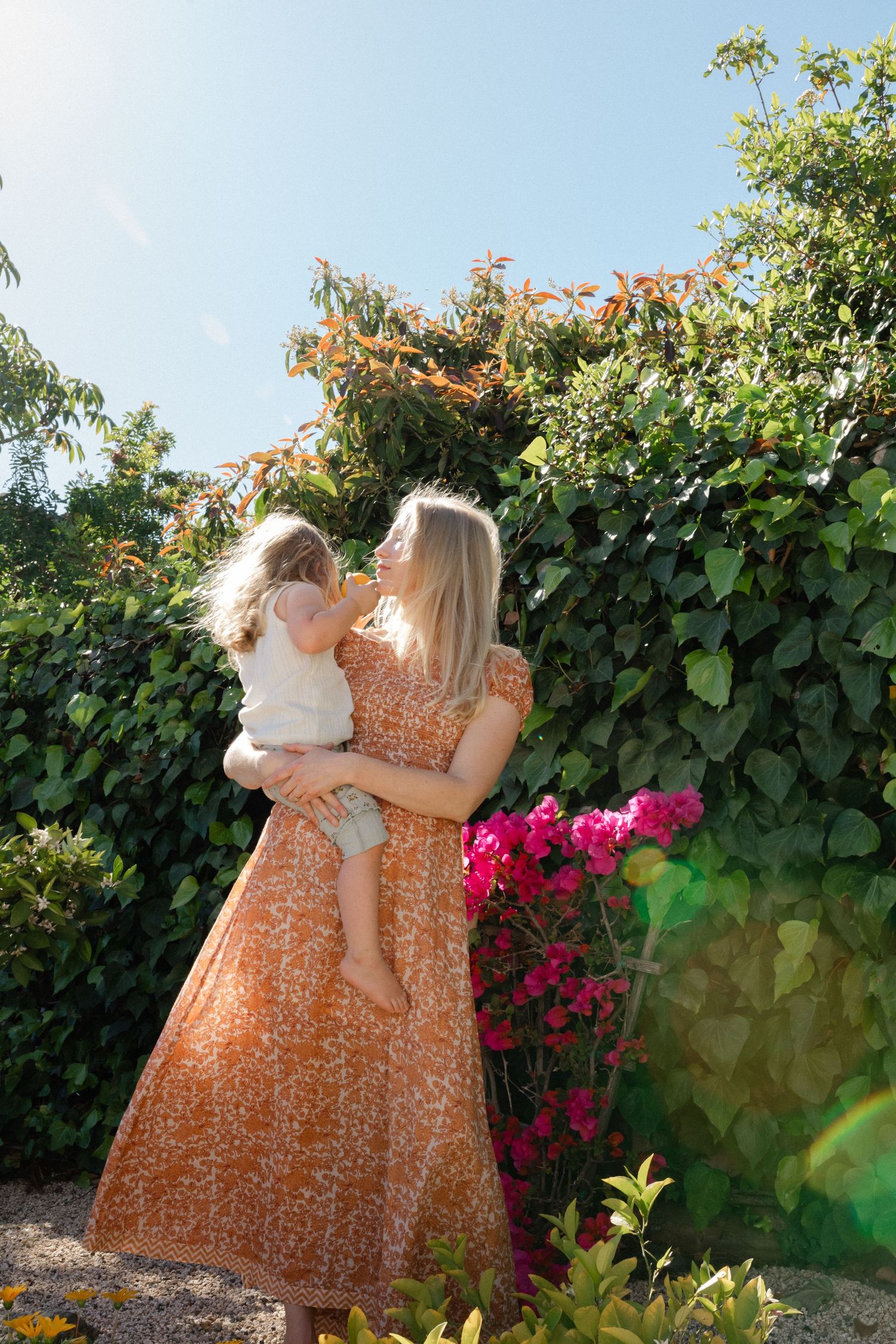 Blonde woman in orange floral dress holds toddler in a lush garden with colorful flowers.