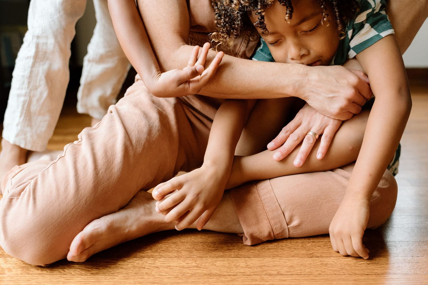 A child sits cross-legged on a wooden floor while an adult gently embraces them from behind.