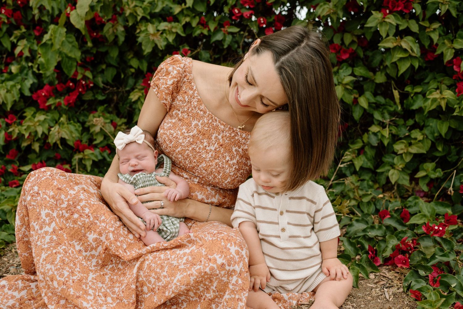 Mother in floral dress sitting outdoors holding newborn baby while toddler sits beside her near red flowers.