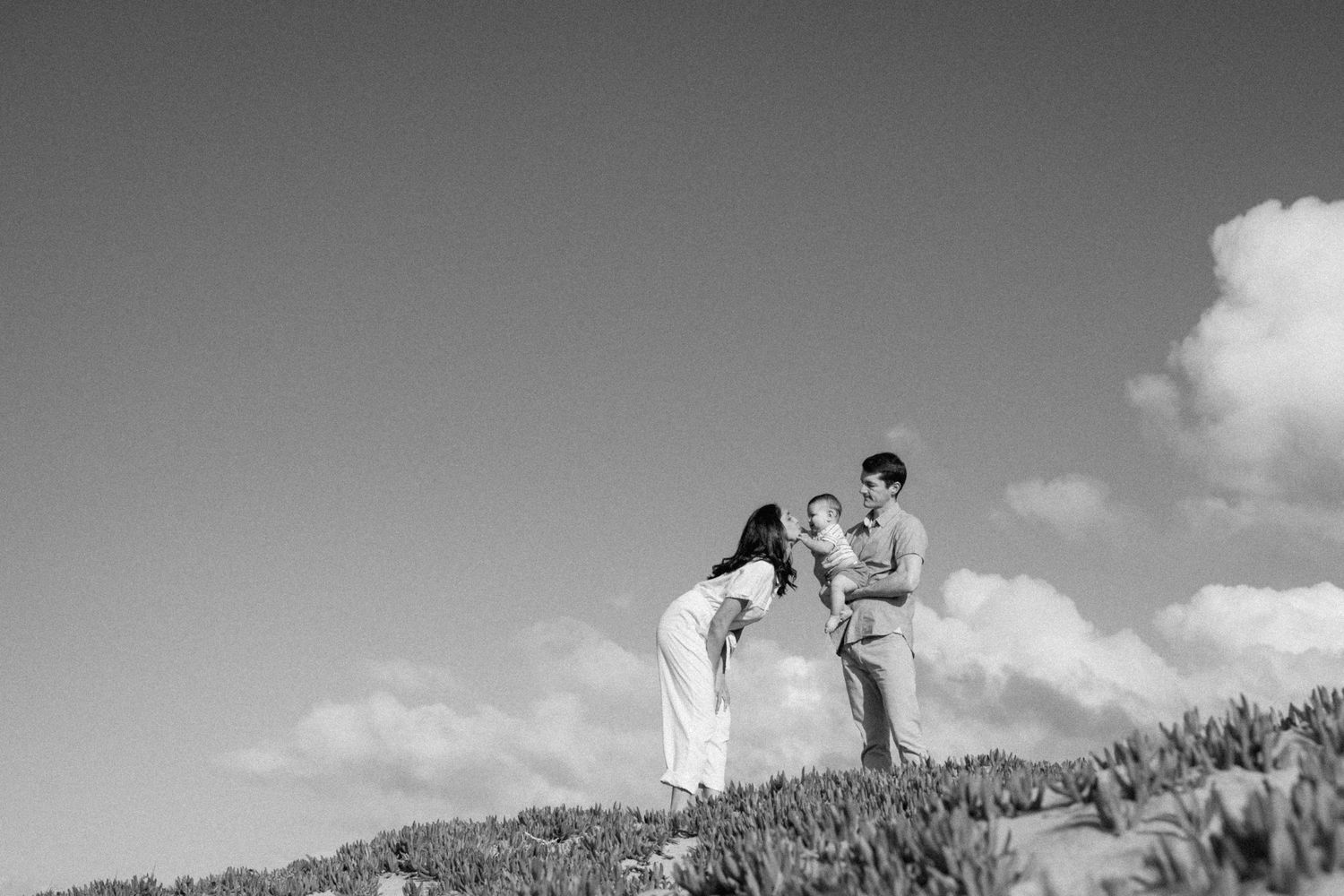 Black and white photo of a couple holding their baby on a hilltop under a cloudy sky.