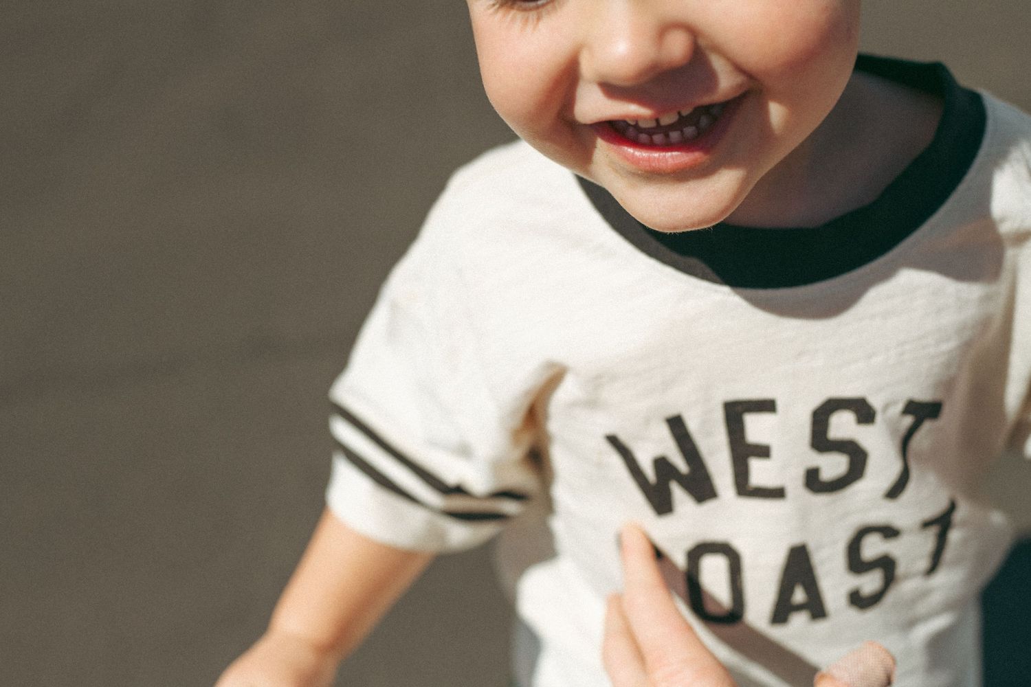Smiling toddler wearing white West Coast graphic t-shirt with black trim outdoors in sunlight.