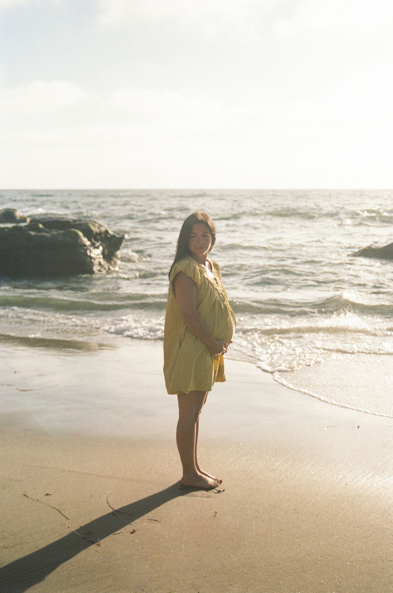 A pregnant woman in a yellow dress stands barefoot on a sunny beach holding her baby bump by the ocean.
