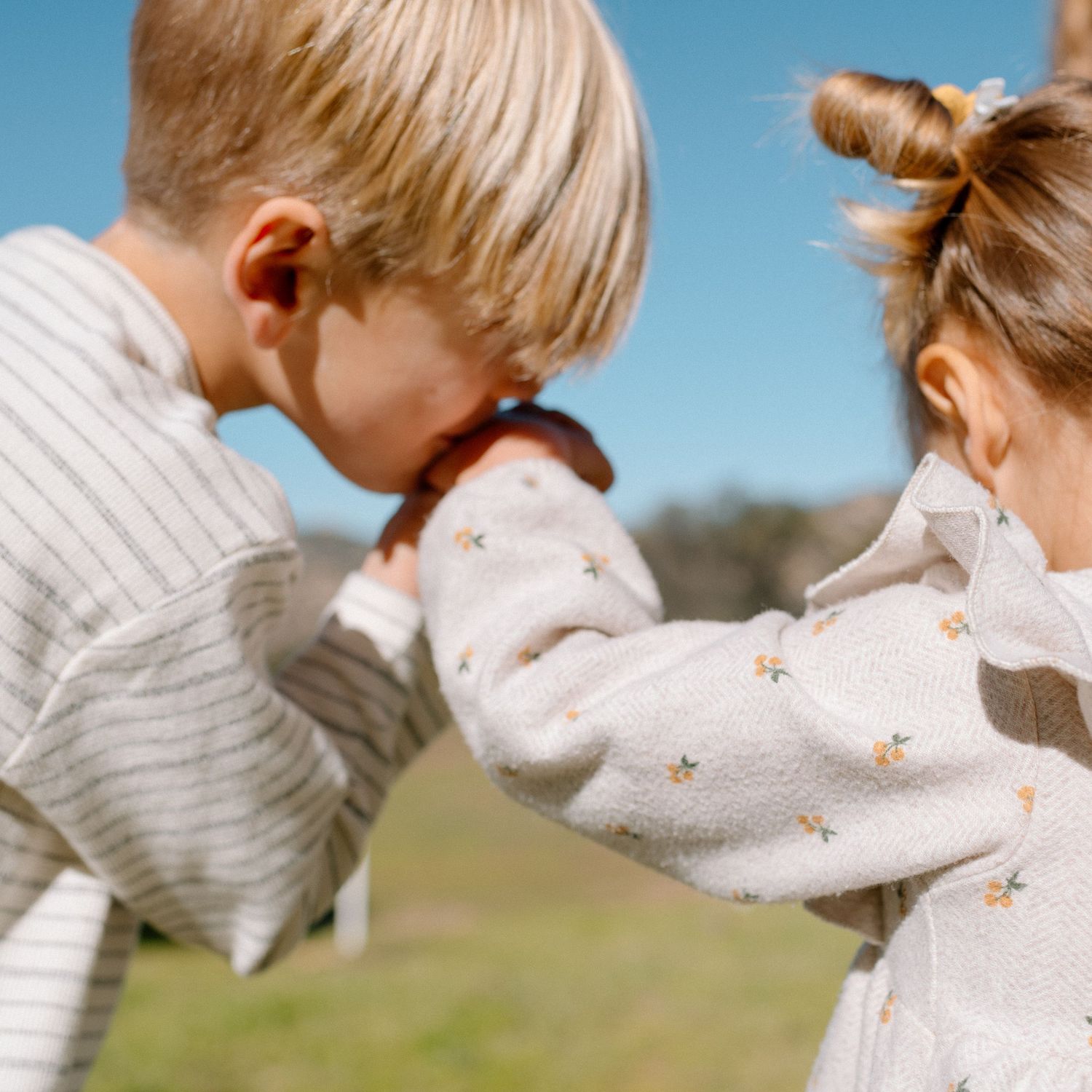 Two young siblings in matching cream sweaters playfully interact outdoors on a sunny day.